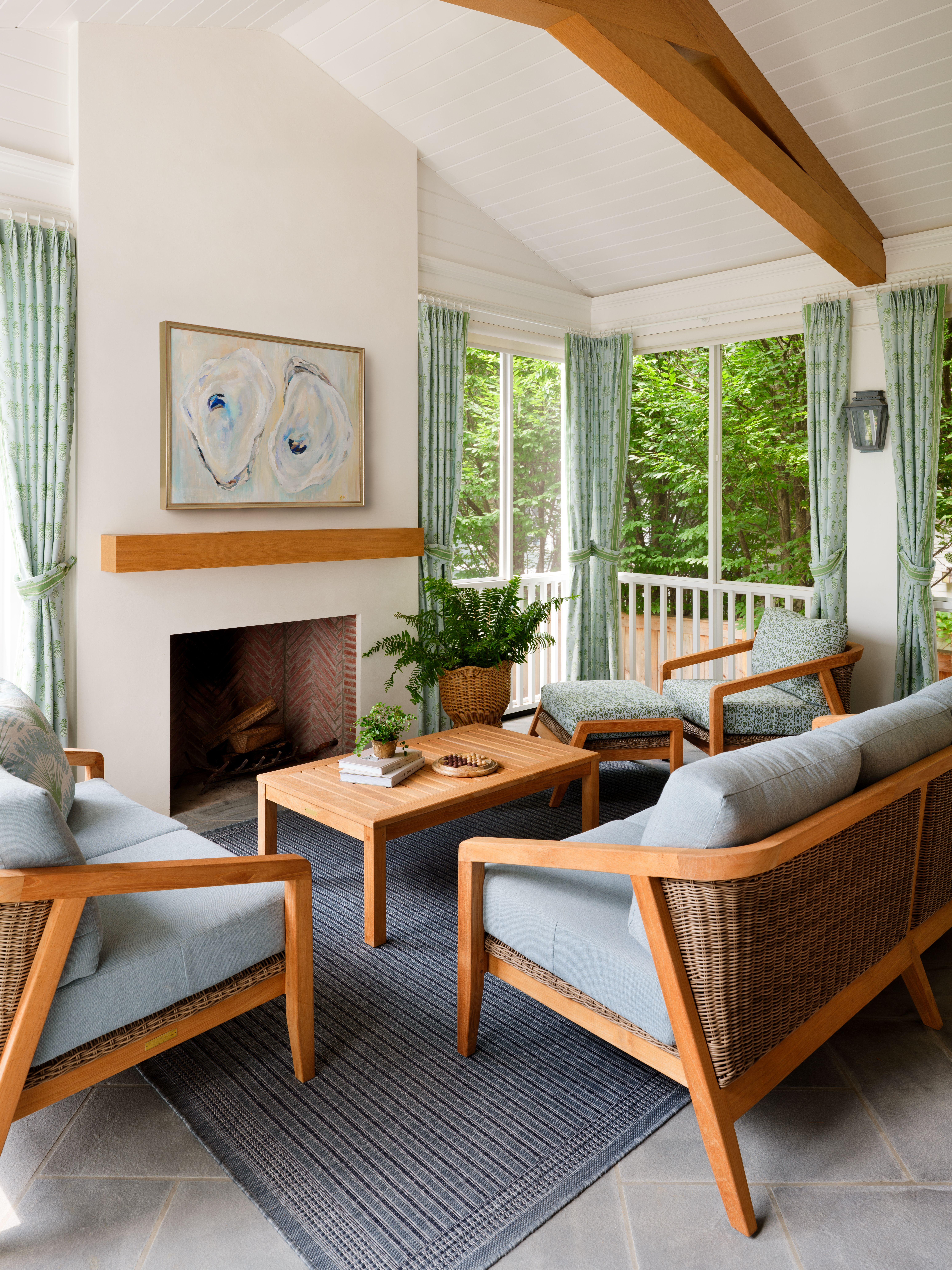 Sunlit porch with white walls and a wood beam ceiling. Wicker chairs with blue cushions around a wooden coffee table, fireplace and green curtains that frame windows to greenery.