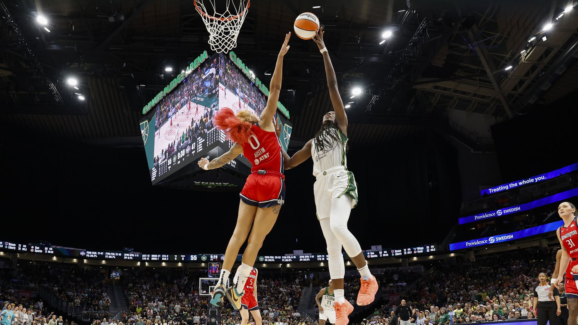 A WNBA player in red seeks to block the shot of another player in white and green. 