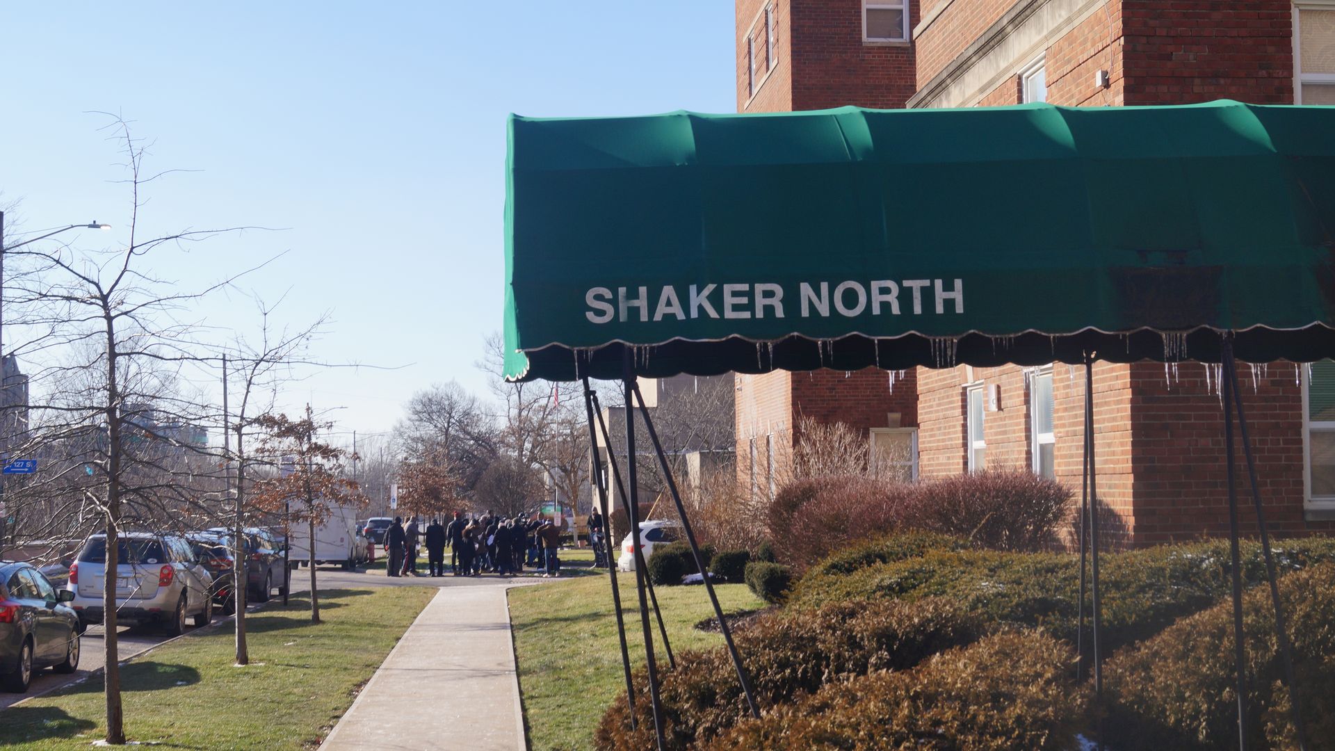 Green awning with "SHAKER NORTH" in white letters over building entrance, sidewalk leading to group of people gathered outside, leafless trees and parked cars on clear sunny day.