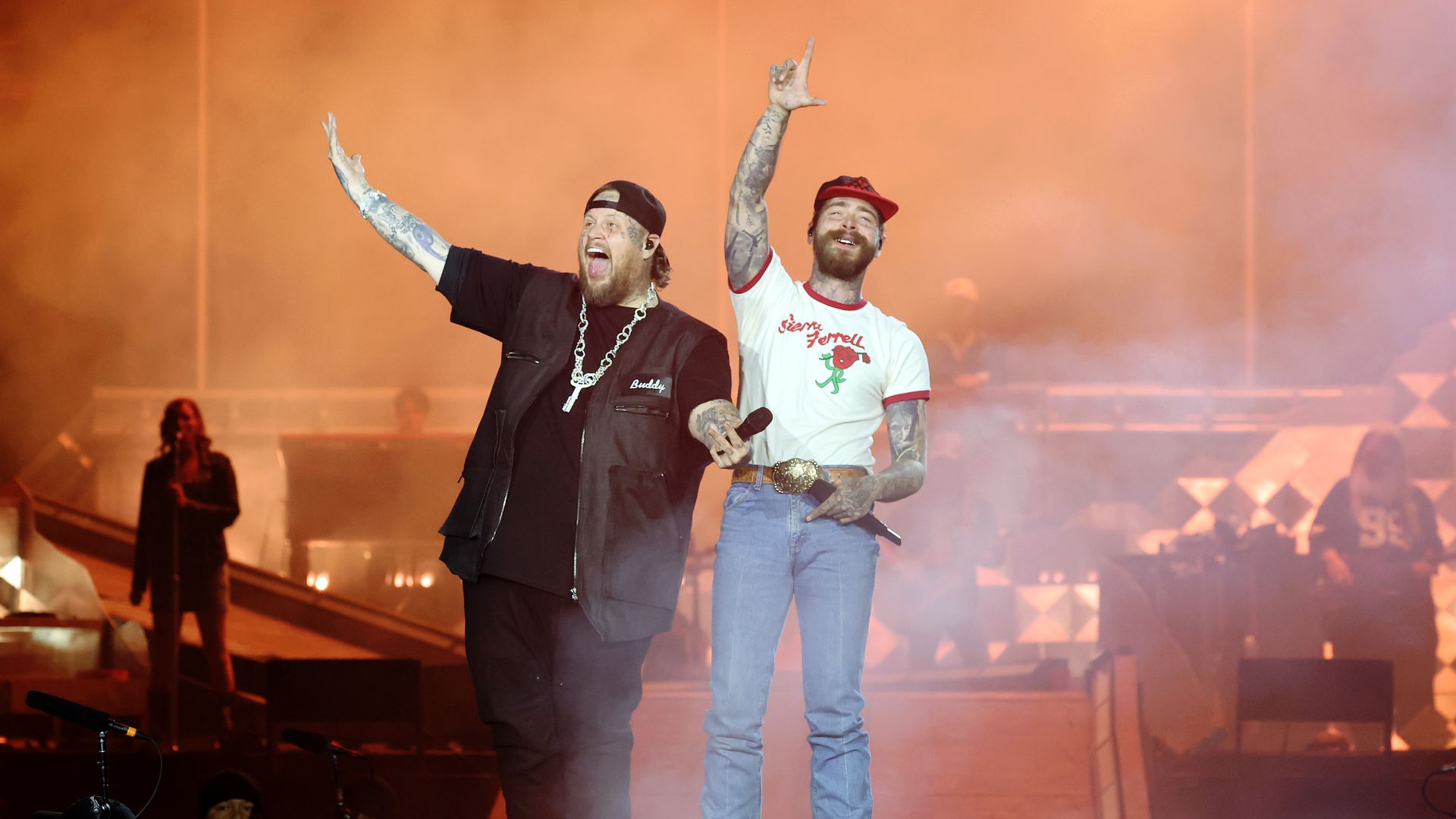 Two tattooed male performers on stage with orange fog, one wearing a black outfit and chain, the other in a white shirt with red trim, blue jeans, and a red cap, both holding microphones.