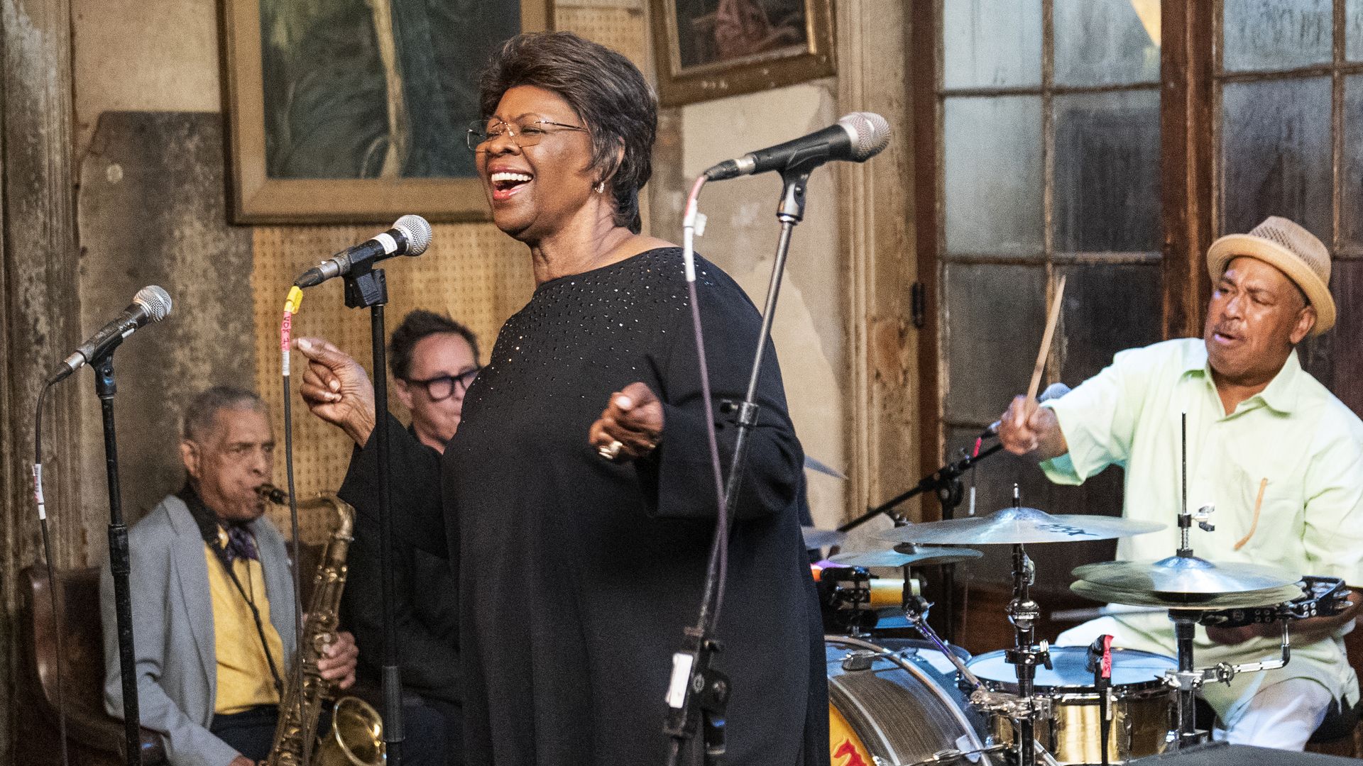 Irma Thomas performs with the Preservation Hall Jazz Band. She sings into a microphone in front of a drummer and musicians playing brass instruments.