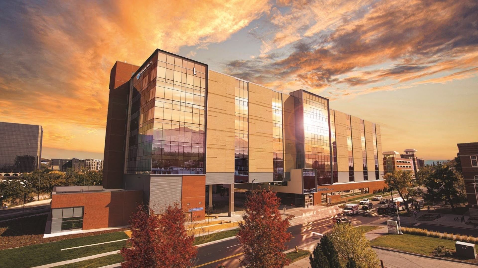 A large modern building with glass panels and brick, bathed in golden sunset light. Reflective windows show an orange sky; street, trees with red leaves, and parked cars line the surrounding campus-like area.