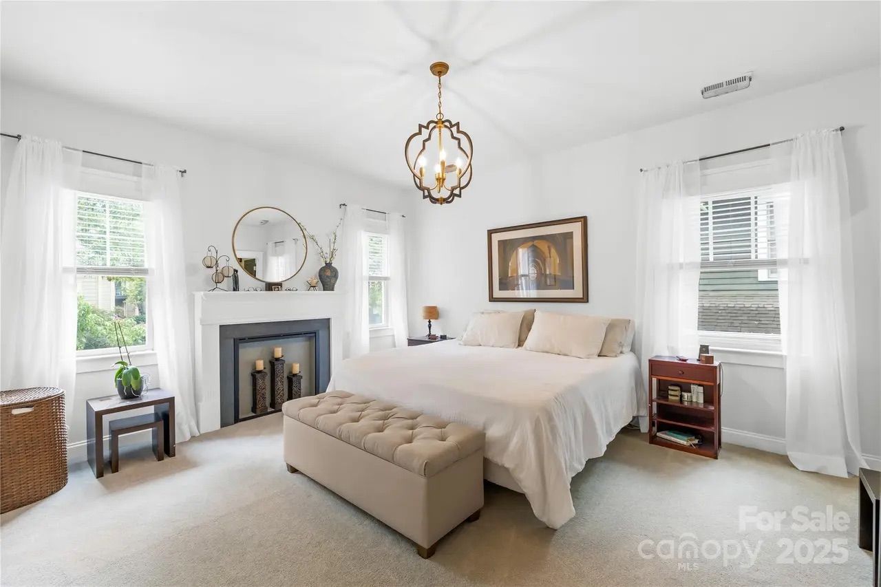 Bright bedroom with white walls, large bed with beige bedding, tufted bench, two windows with sheer curtains, a fireplace with candles, round mirror, and a decorative chandelier.
