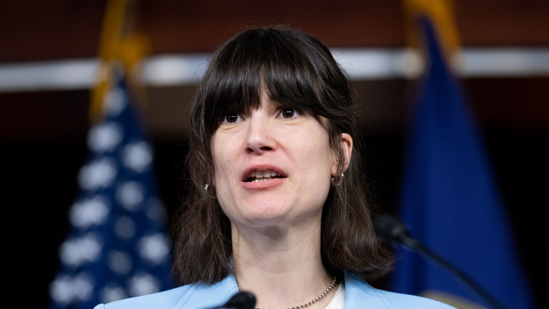 A young US lawmaker with dark hair and a blue outfit stands at a podium in front of flags.