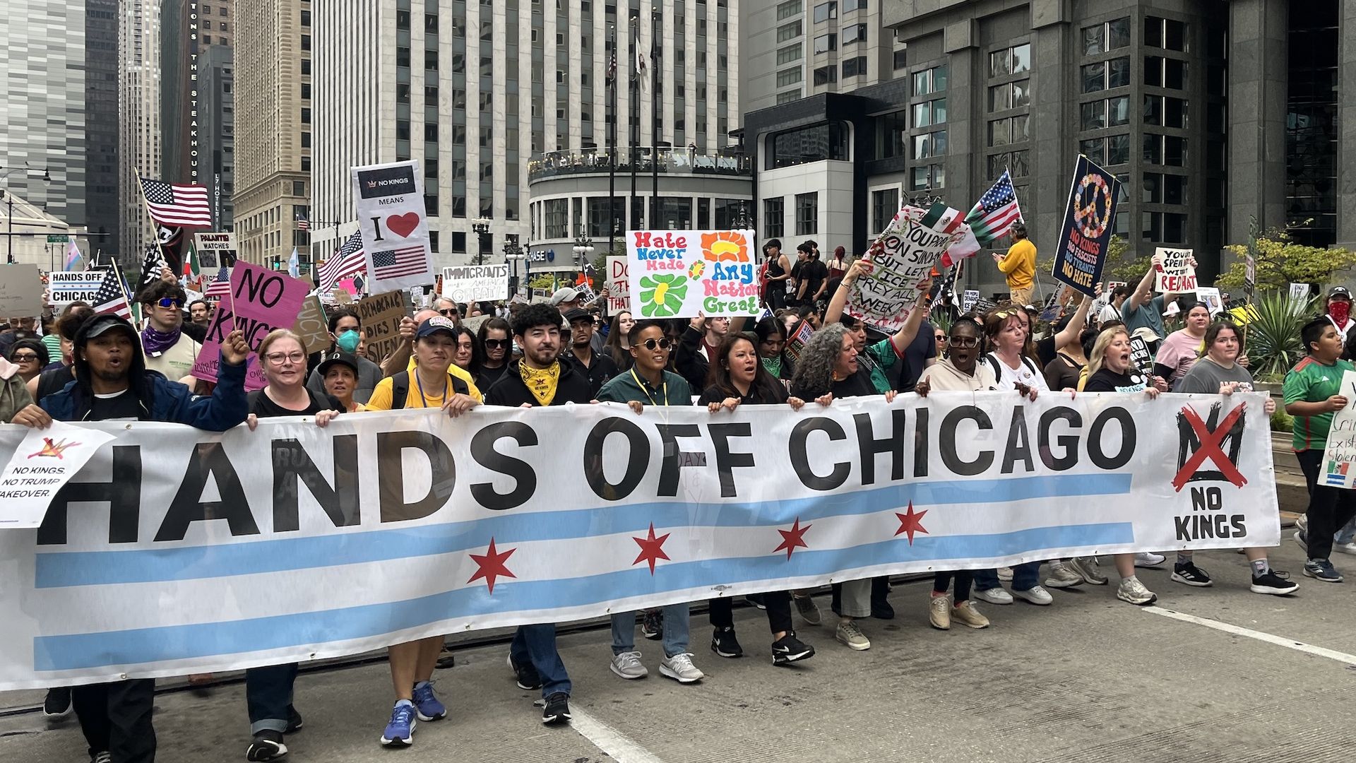 Diverse protesters march through a city street, holding a long banner reading "HANDS OFF CHICAGO" with blue stripes and red stars; many signs and flags rise as tall skyscrapers loom behind them.