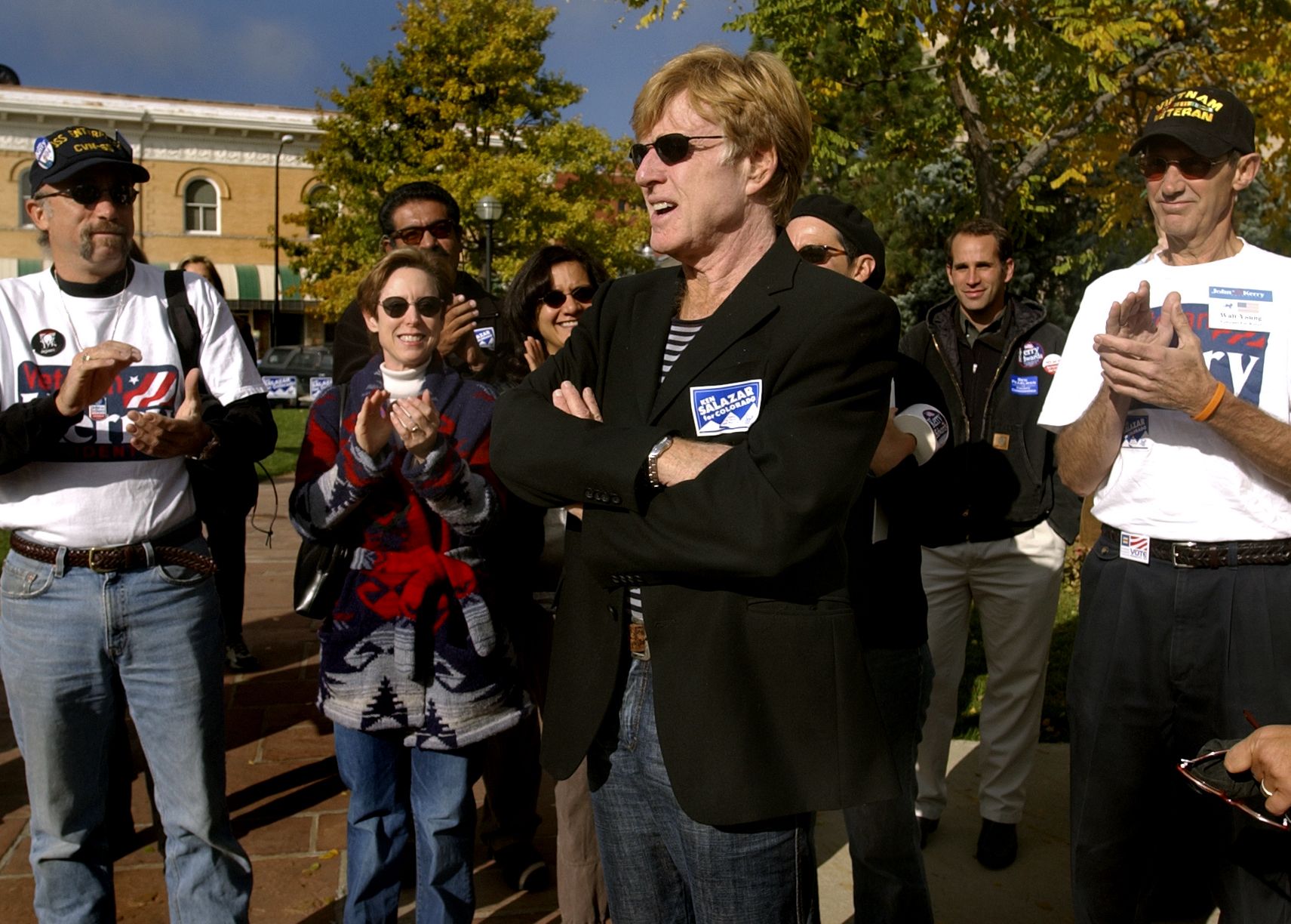 Robert Redford, in a jacket and sunglasses, is surrounded by an outdoor gathering of people clapping and smiling.