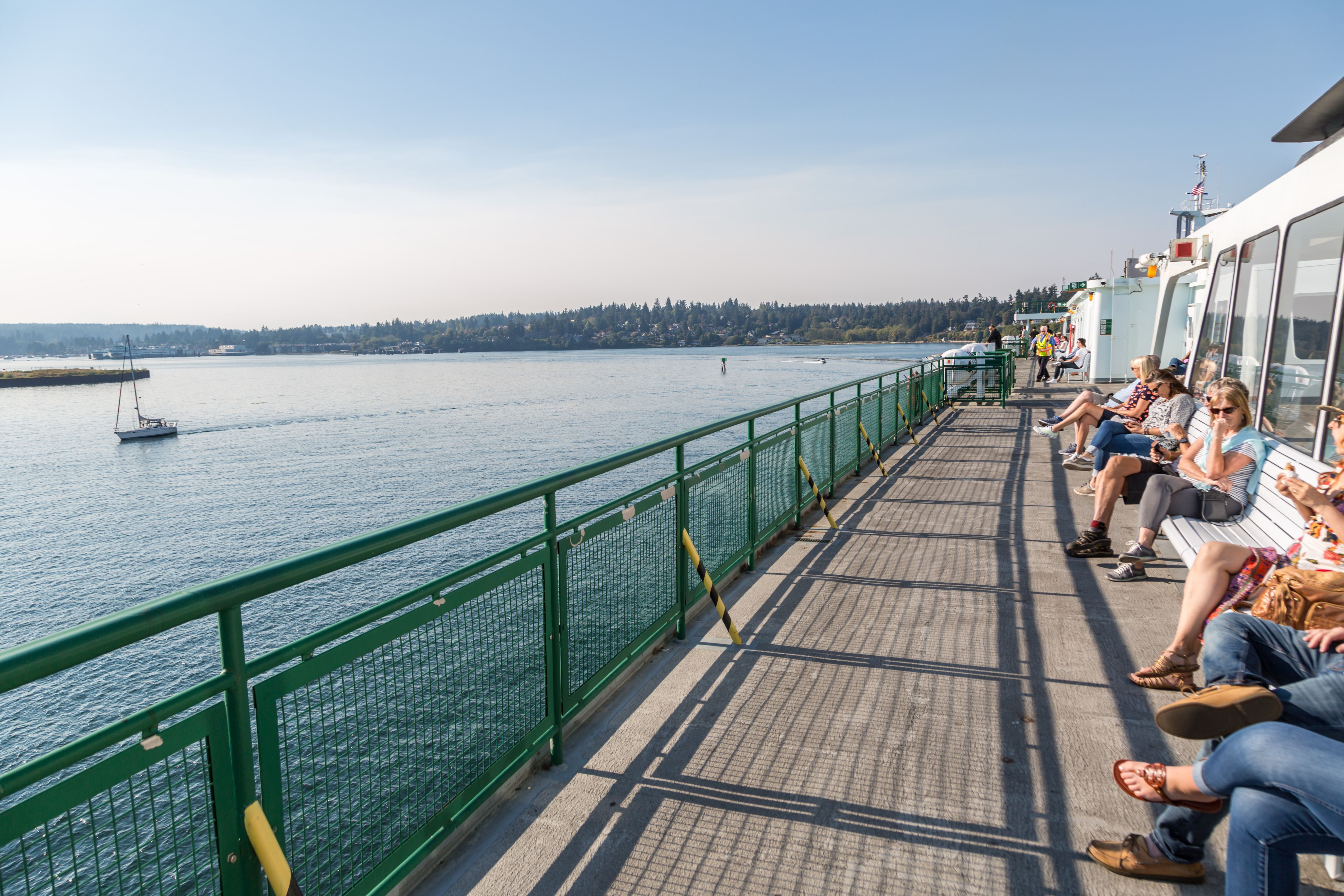 People sitting and relaxing on benches along the green railing of a ferry deck overlooking calm blue water and a sailboat under a clear sky.