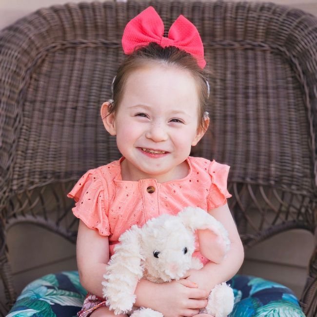 A young girl with a pink bow sitting on a wicker chair.