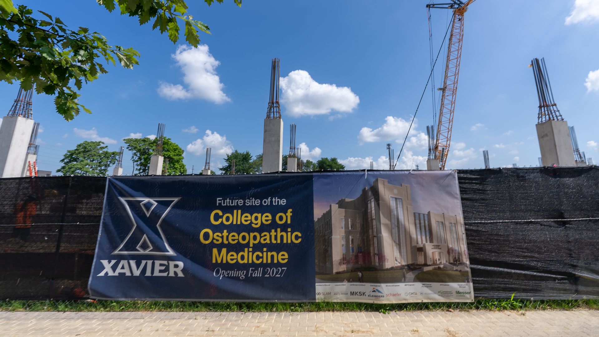 Blue-sky construction site with an orange crane on the right, concrete pillars, and a banner reading "Future site of the College of Osteopathic Medicine" at Xavier, framed by green tree branches.