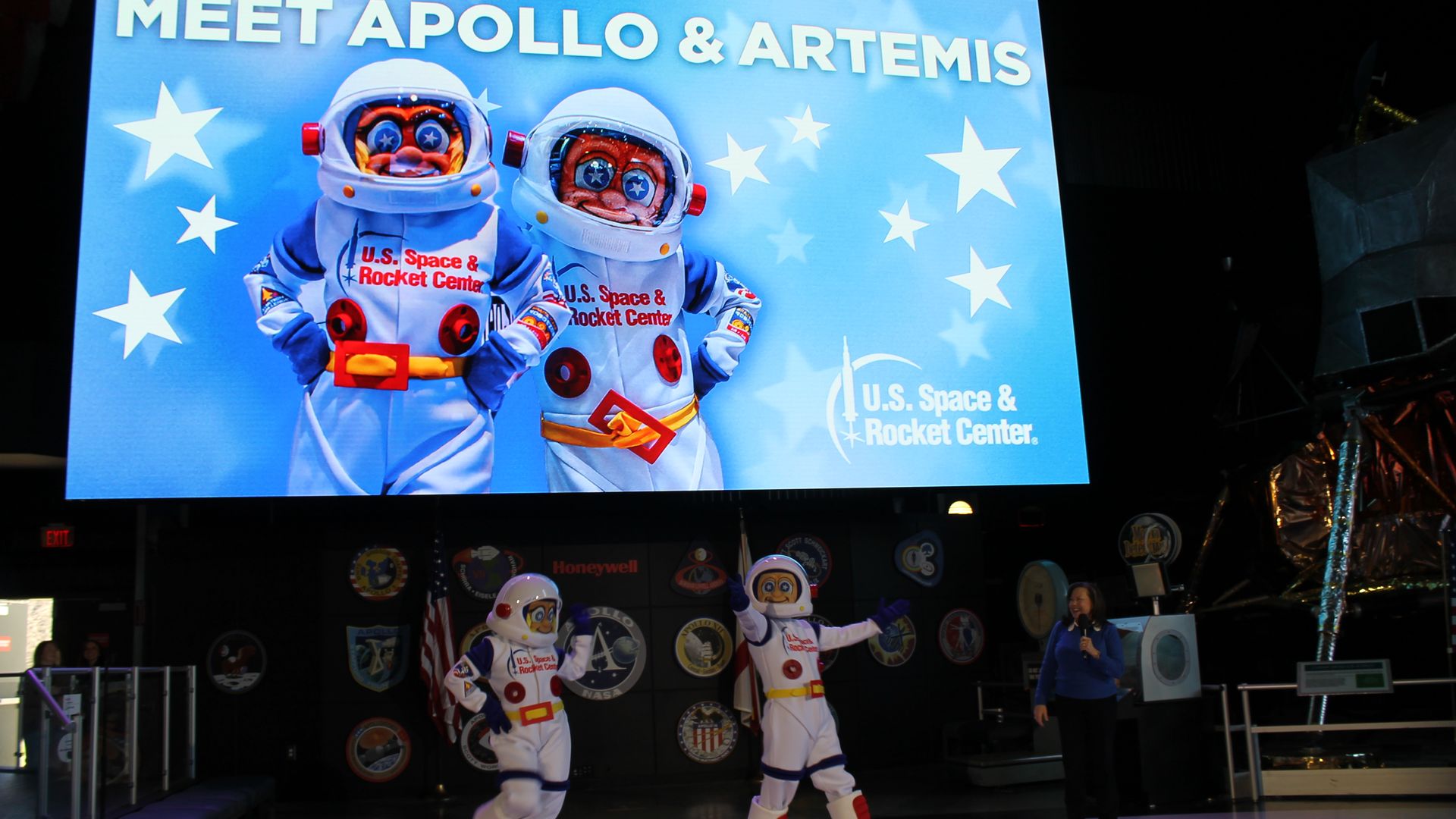 Blue display reads "MEET APOLLO & ARTEMIS" with two astronauts in white suits. In front, two costumed figures walk on a purple floor as a presenter with a microphone stands to the right.