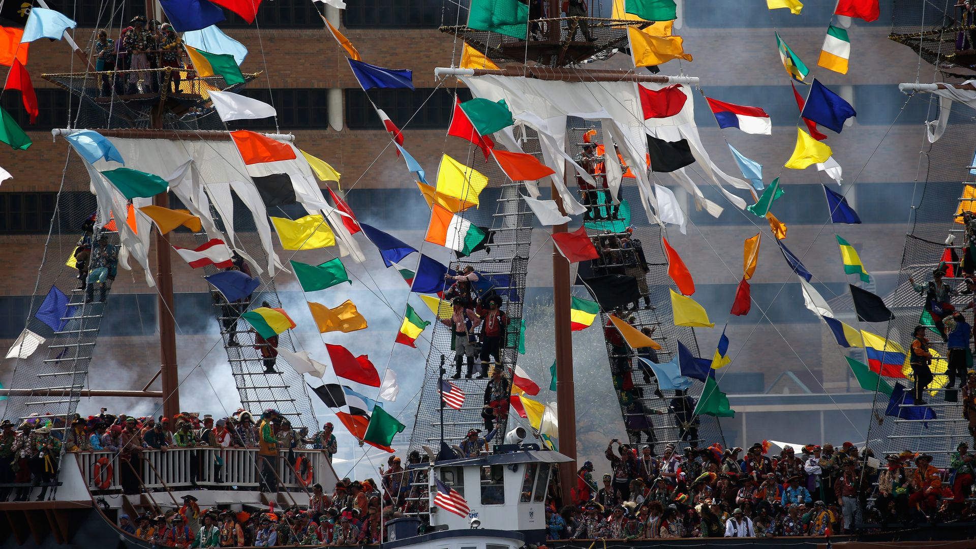 Crowd of people dressed as pirates on a large ship decorated with colorful maritime signal flags against a city building backdrop, with some people climbing the rigging.