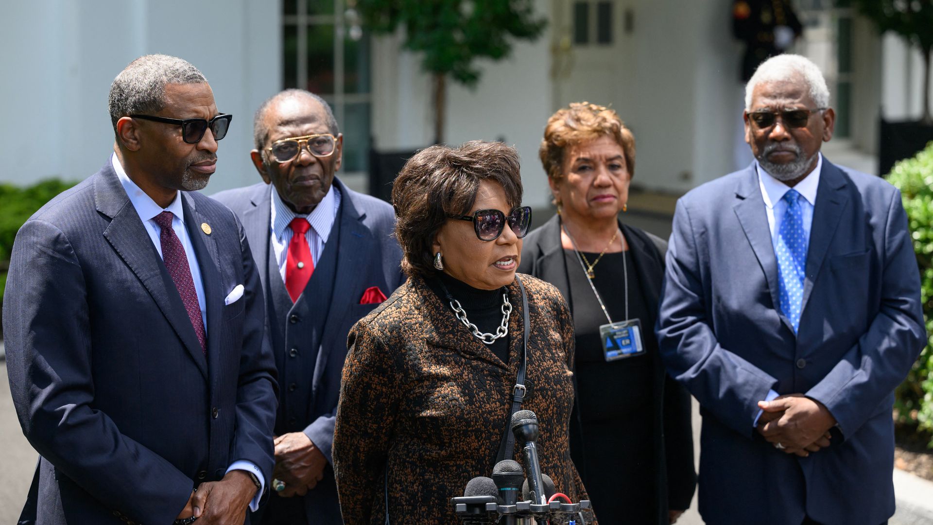 Five people stand outdoors with one speaking into several microphones.