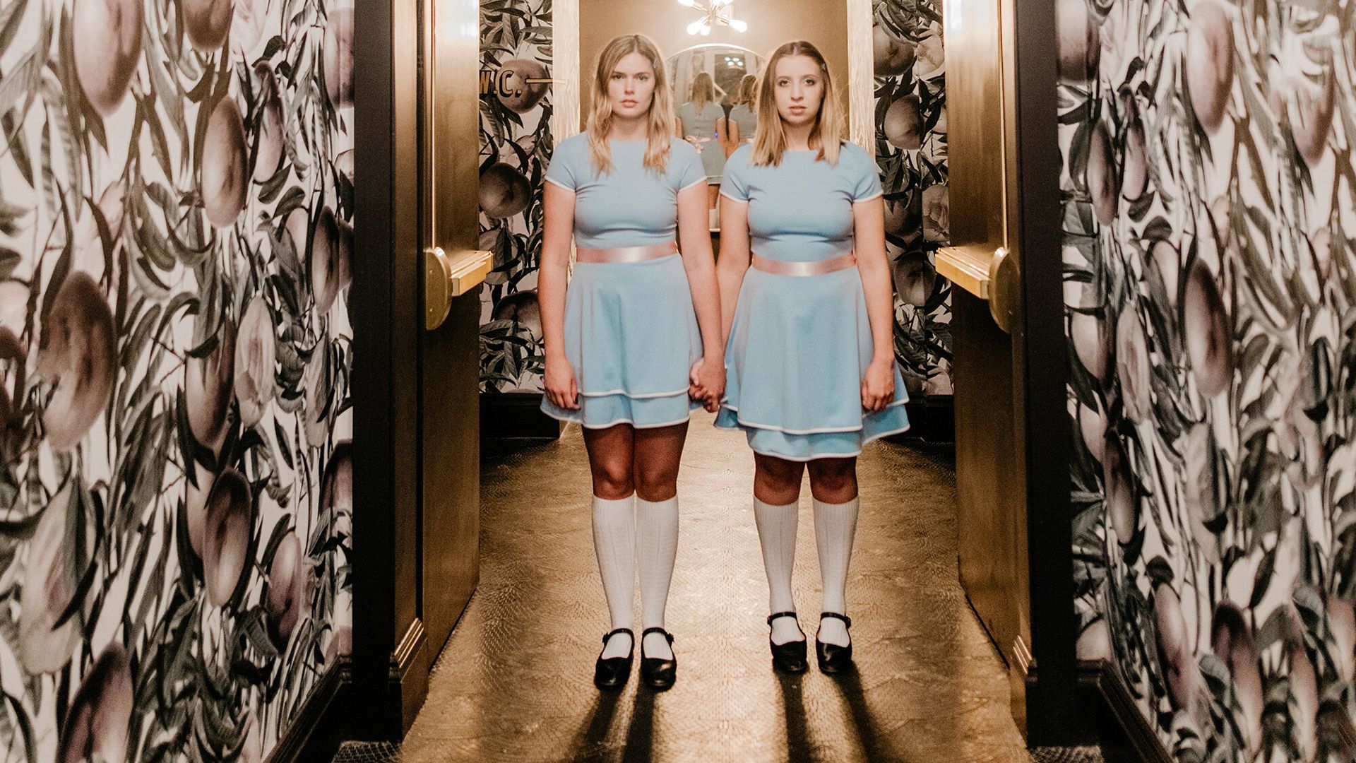 A photo of two blond women recreating the iconic scene of two twins standing in a long hallway in The Shining. 