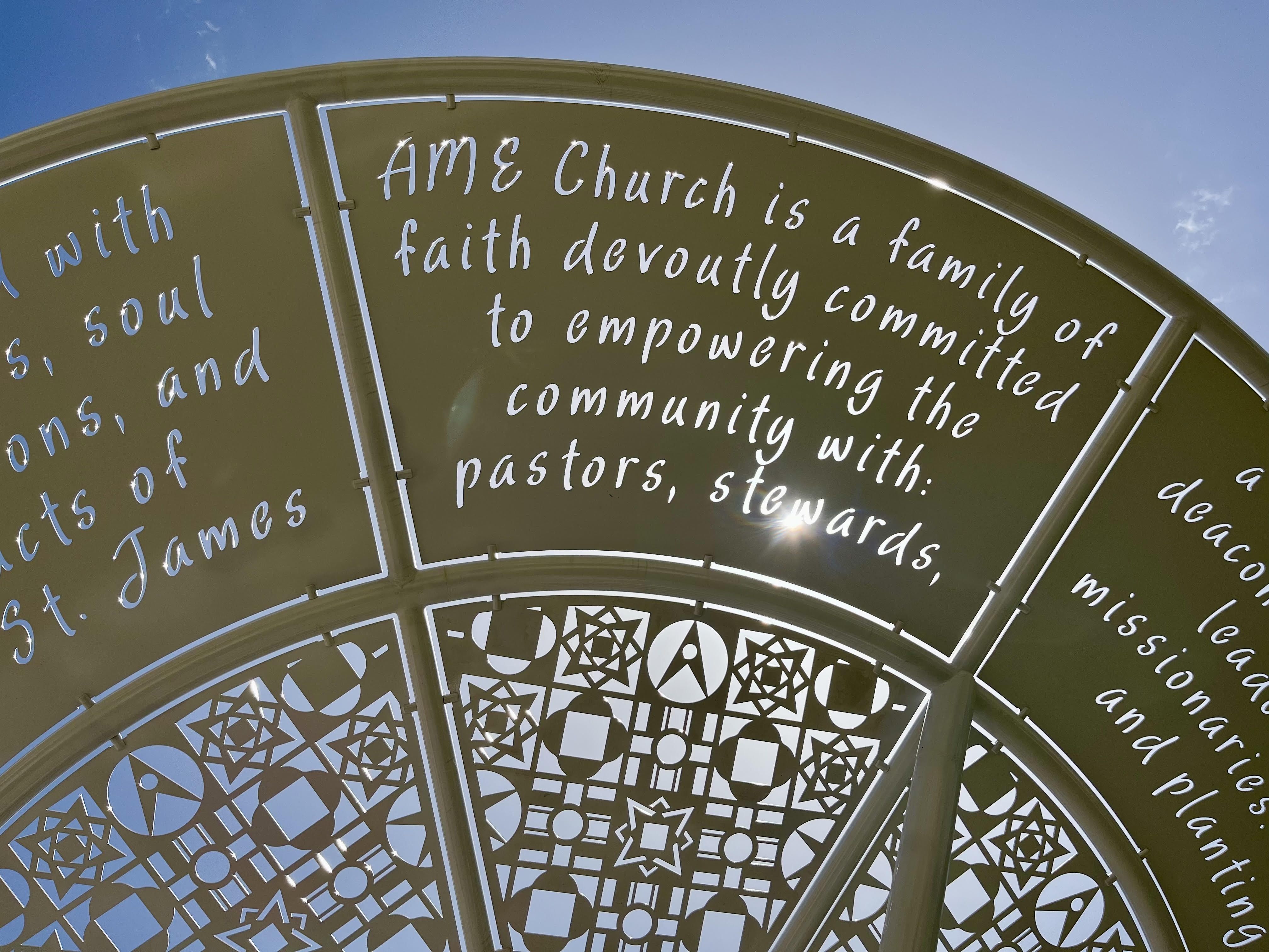 A close-up of an arched pavilion with text cut against the sky that reads "AME church is a family of faith devoutly committed to empowering the community with pastors, stewards." Etched panels beneath the text show quilt patterns.