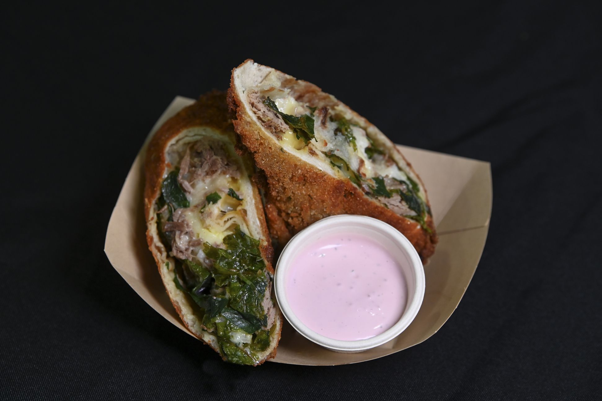 Close-up of a fried sandwich cut in half, stuffed with shredded meat, wilted greens, and melted cheese, served in a brown paper tray with a pink dipping sauce on a dark background.