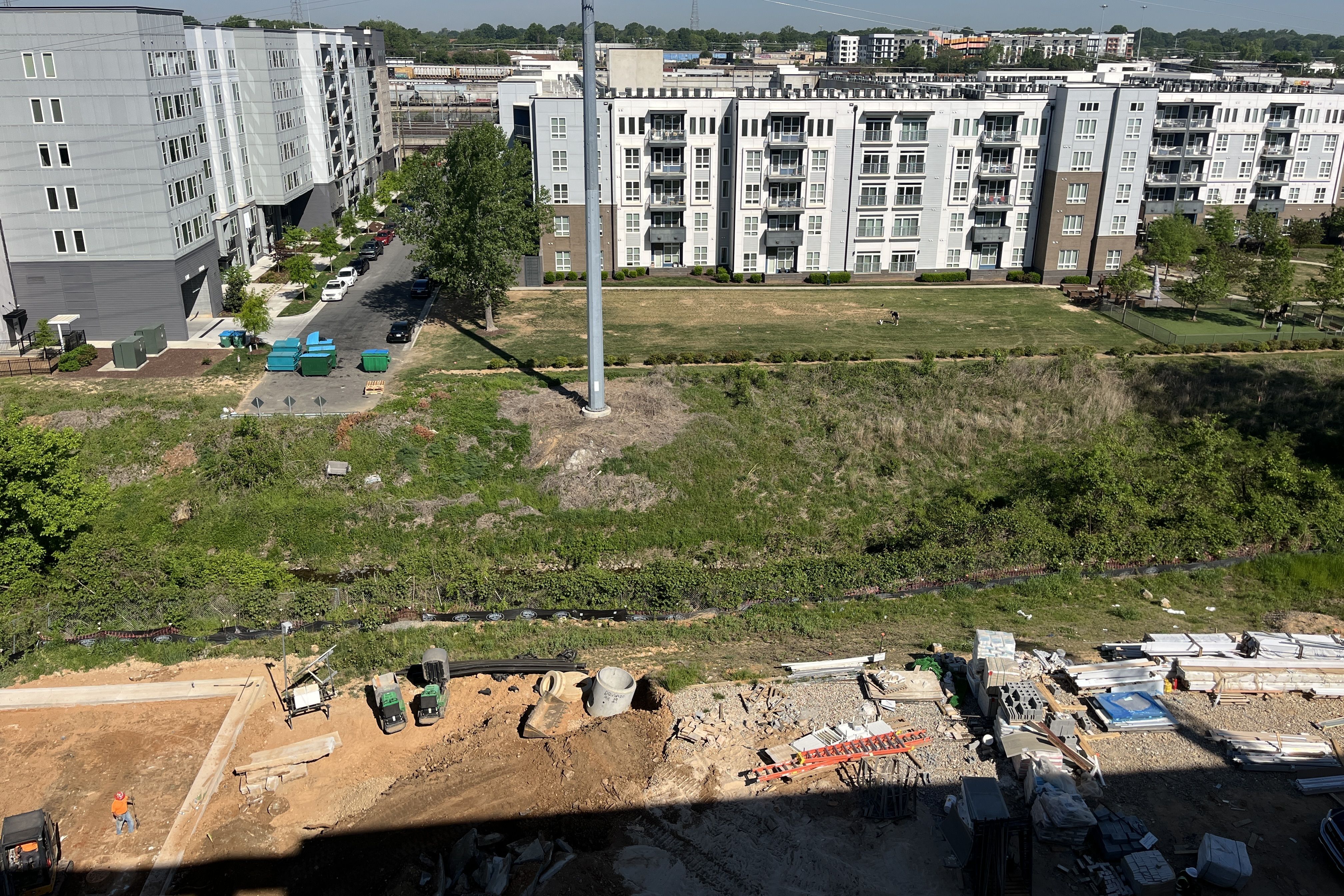 Aerial view of a modern apartment complex with gray multi-story buildings and balconies, a street between them, and a foreground construction site with dirt, pallets, and an orange-vested worker.