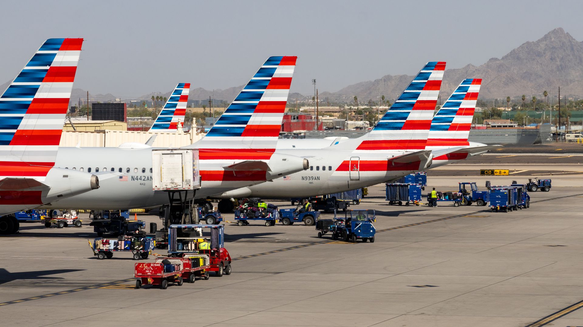 Five American Airlines planes lined up on the tarmac with red, white, and blue striped tails, surrounded by blue and red baggage carts and workers under a clear sky.