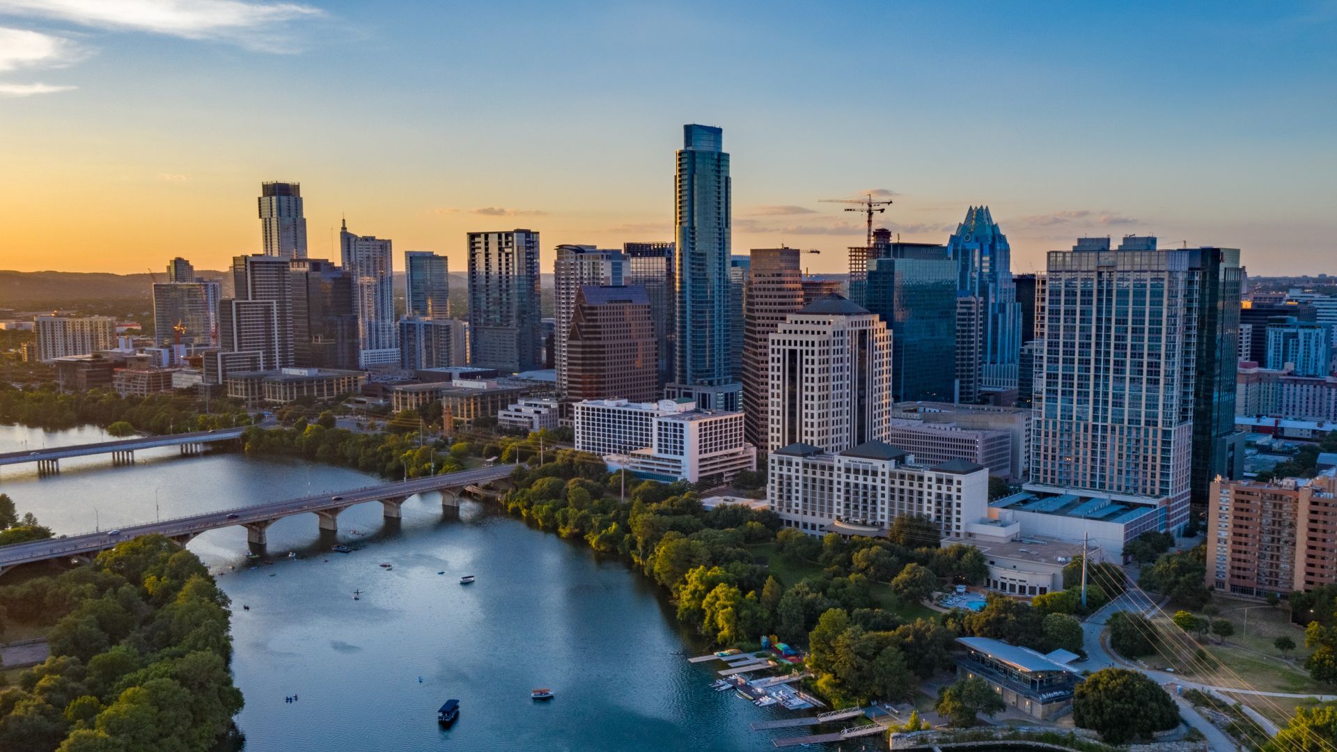 Panoramic city skyline at sunset: glass skyscrapers, a river with a bridge, and green banks. Orange sky fades to blue; construction cranes dot the skyline.