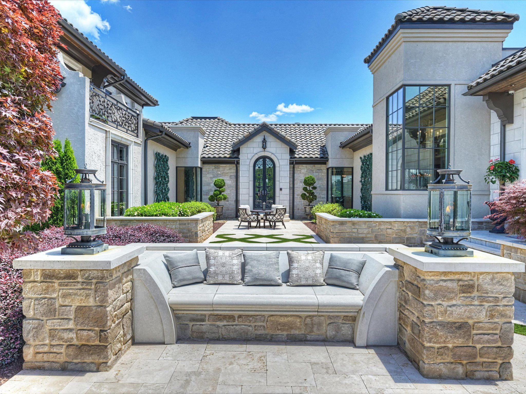 Stone-built outdoor seating area with gray cushions, flanked by two lanterns, in a courtyard with trimmed bushes and a table with chairs in front of a modern beige house under a blue sky.