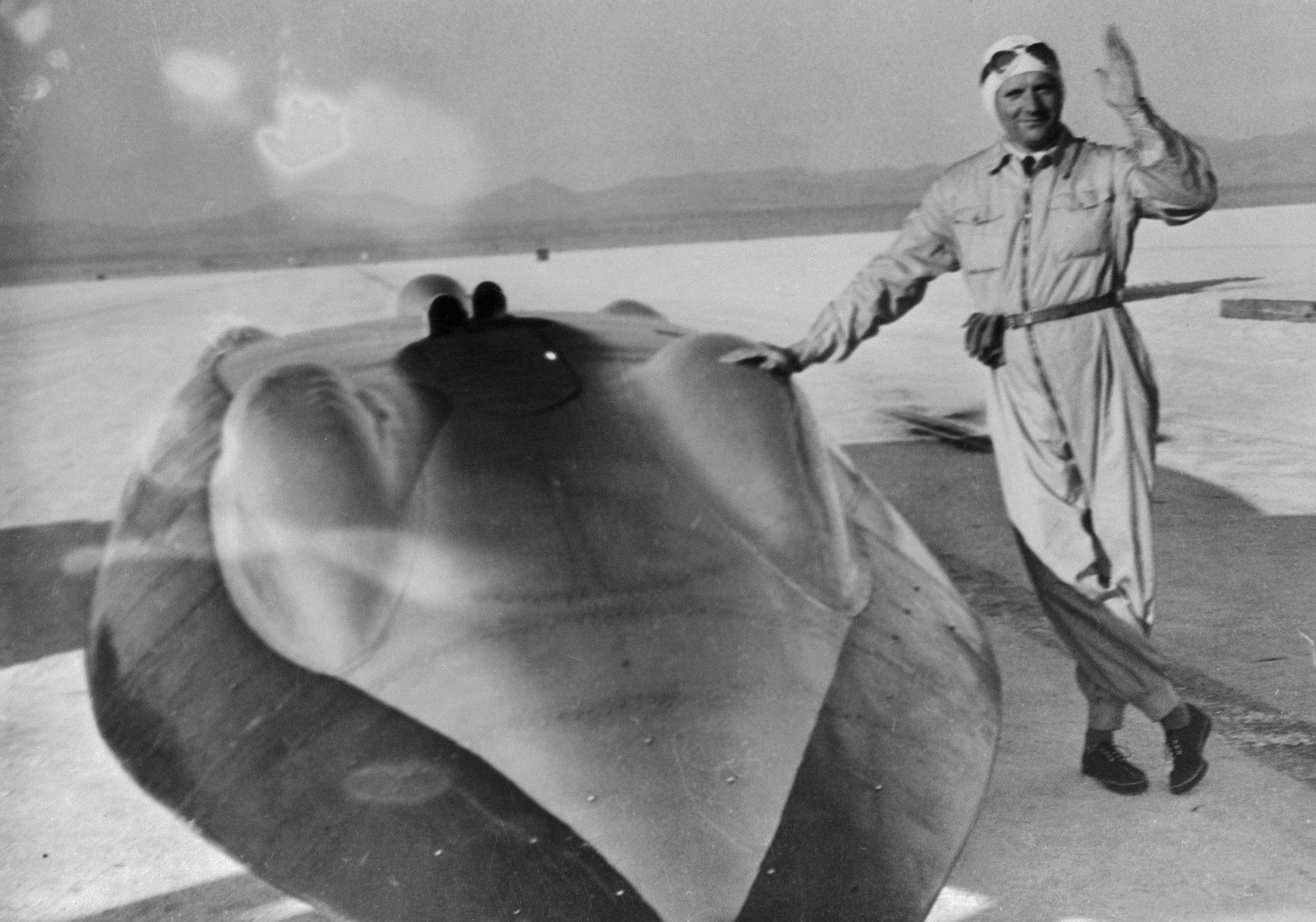 A driver in coveralls and goggles stands next to a race car in the 1930s on salt flats