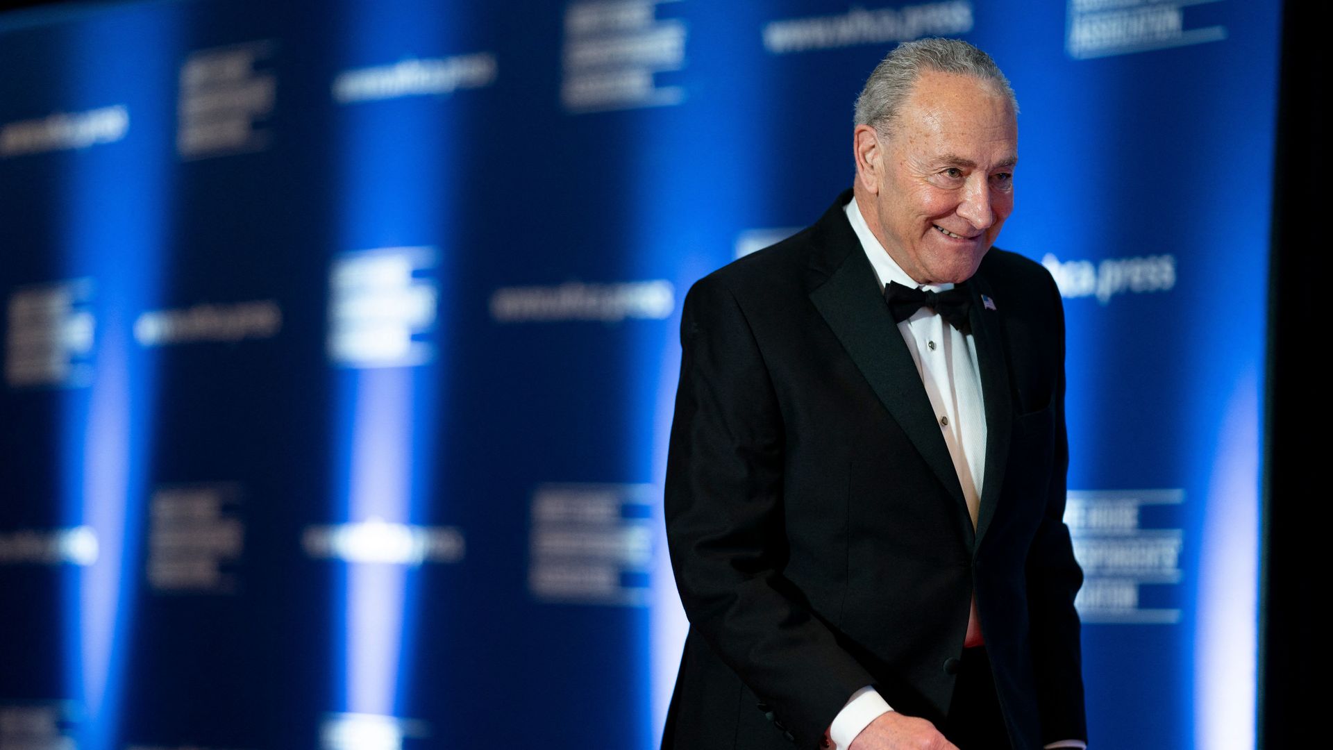 Chuck Schumer, wearing a bowtie in front of a blue "White House Correspondents Association" backdrop, walks as he is photographed at the White House Correspondents Association dinner.
