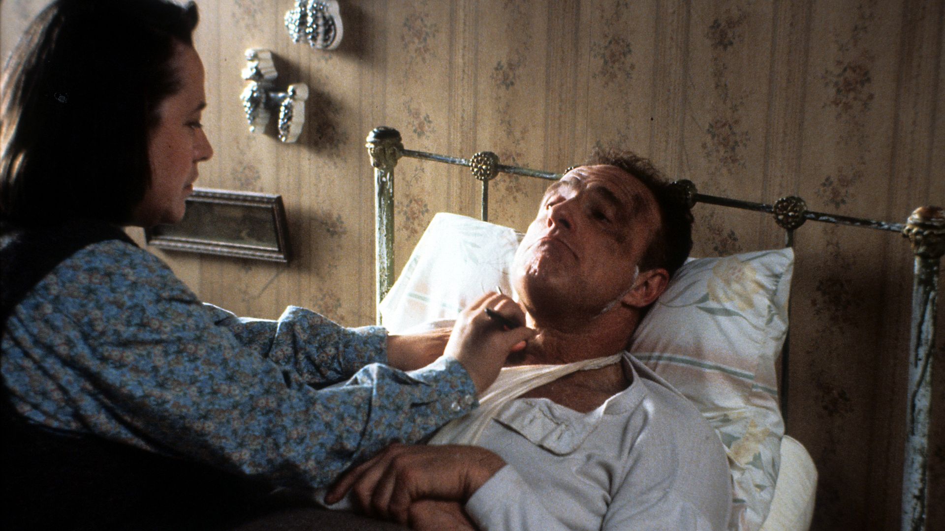 A woman in a blue floral shirt tending to a man with a black eye lying in bed with an old metal frame, beige wallpaper, and floral pillows in a dimly lit room.