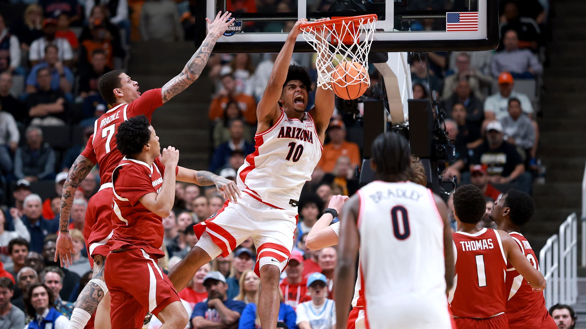 A University of Arizona basketball player in a white uniform yells while dunking the basketball in a packed arena. He's surrounded by opponents in red uniforms while a teammate with his back to the camera looks on. 