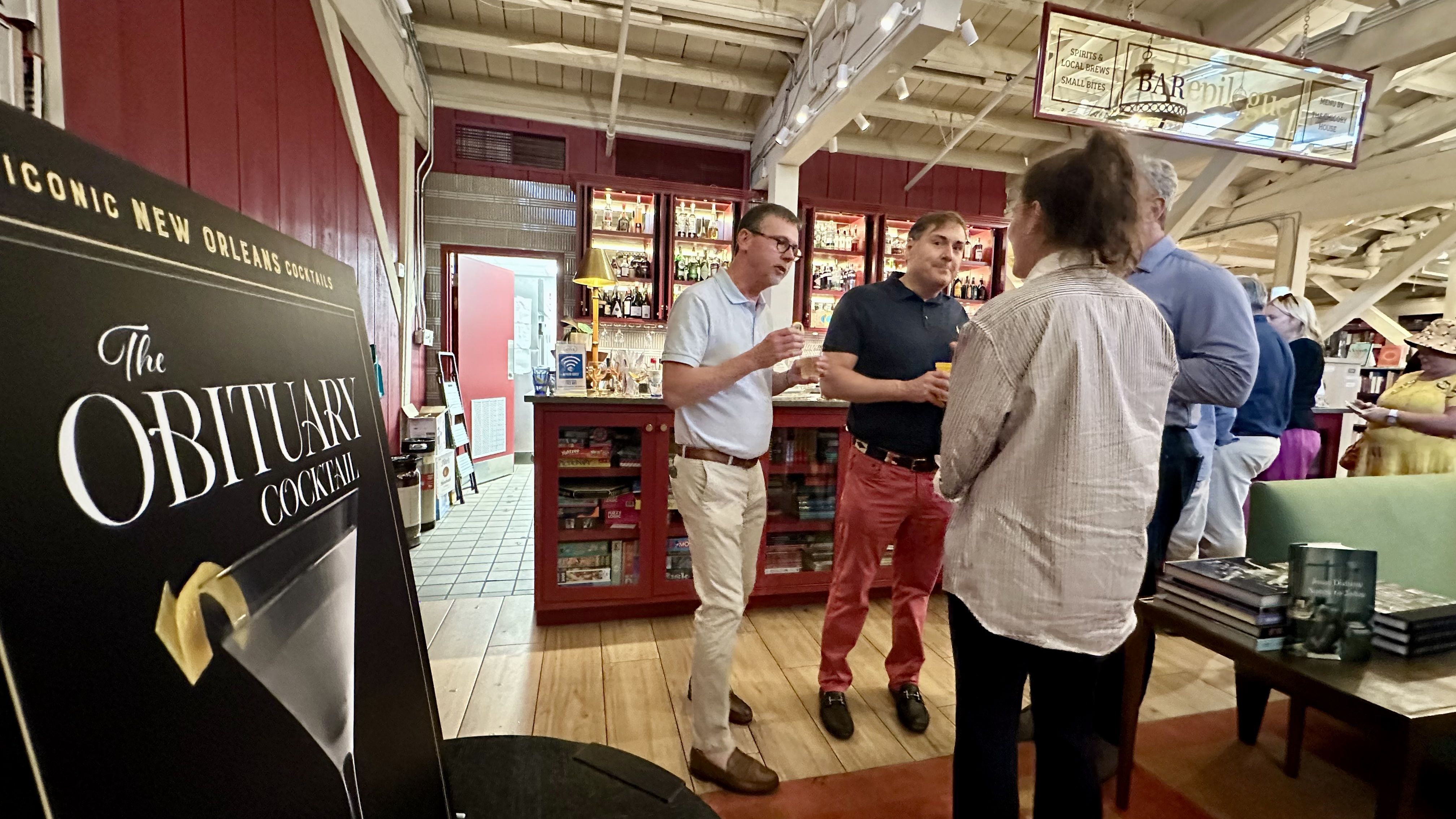 People socializing in a warmly lit bar with red walls and wooden floors, near a sign advertising "The Obituary Cocktail," an iconic New Orleans drink, with shelves of bottles and books nearby.