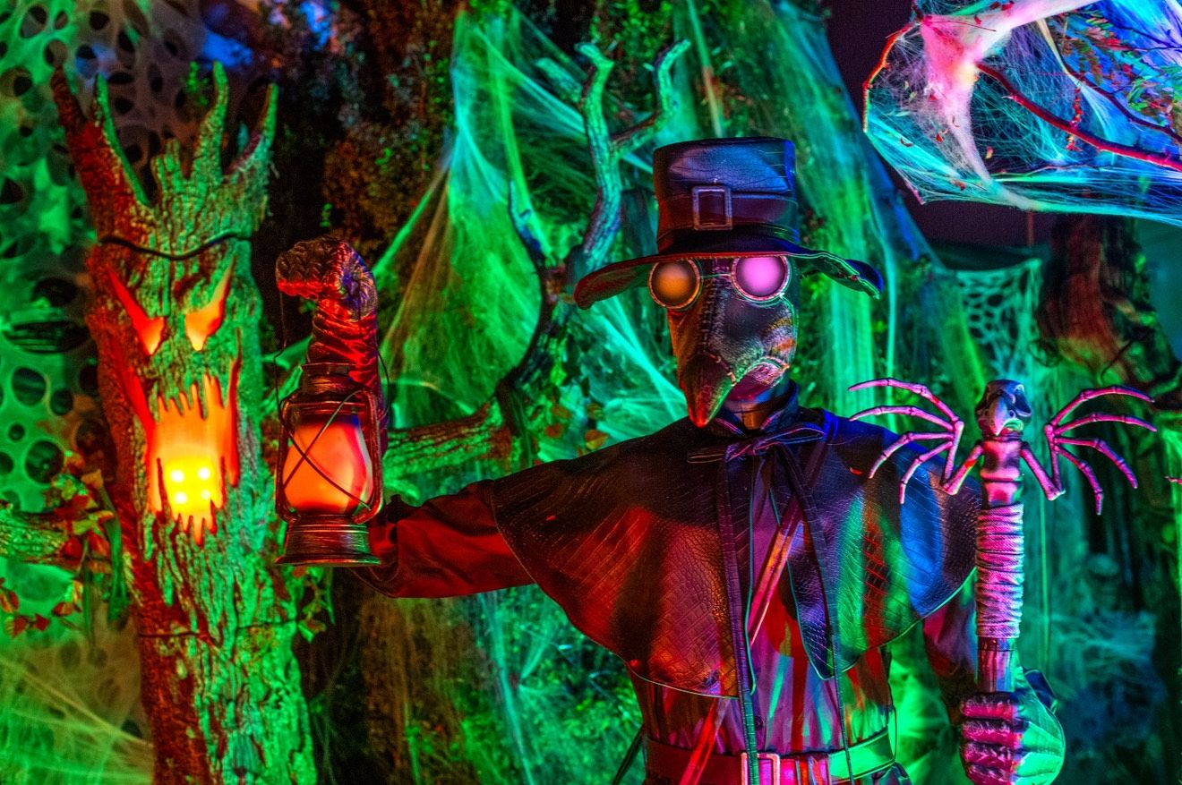 A figure in a plague mask holding a lantern surrounded by haunted features and spider webs