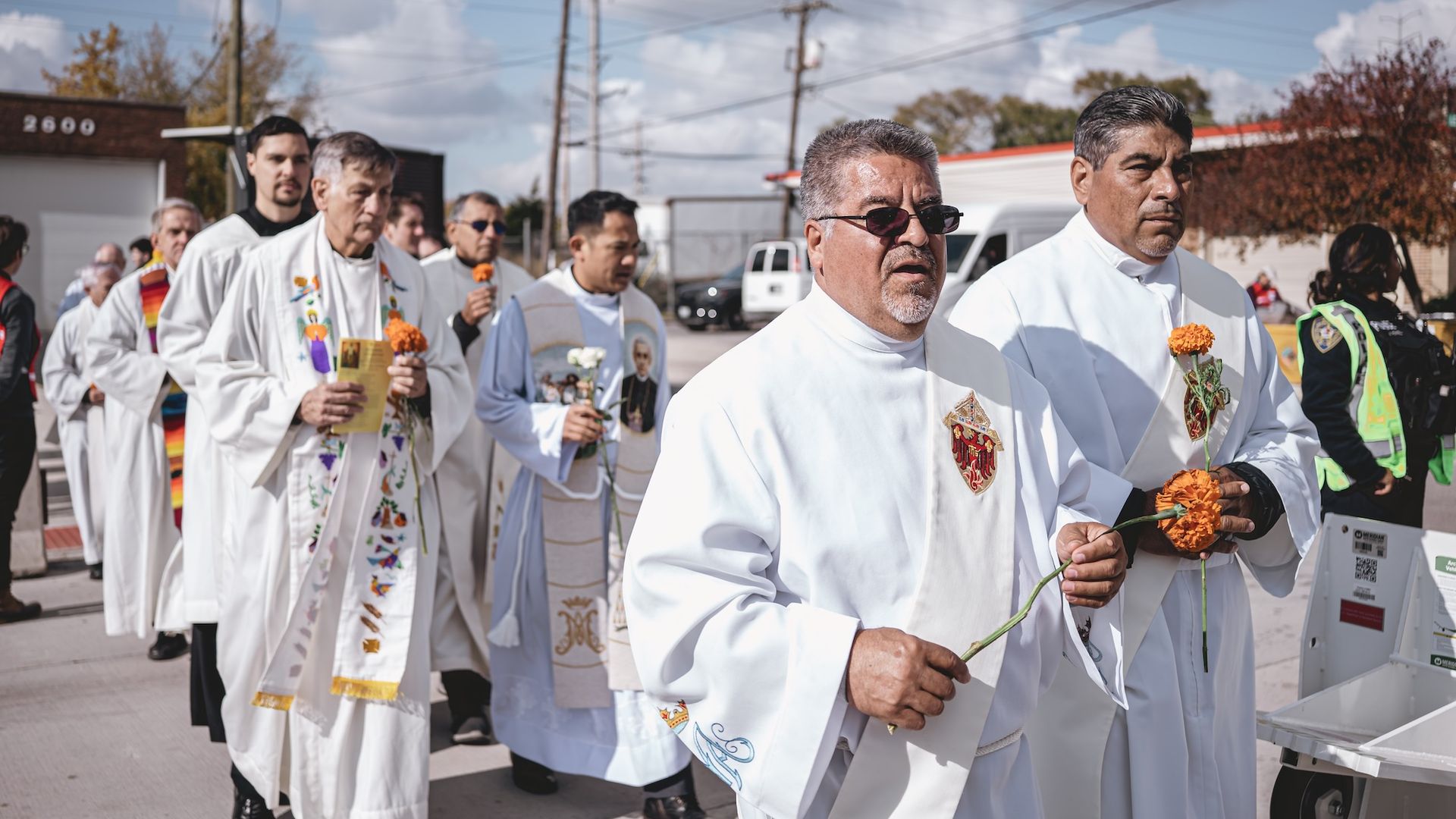 Group of men in white religious robes holding orange and white flowers, walking outdoors on a sunny day with blue sky and power lines in the background.