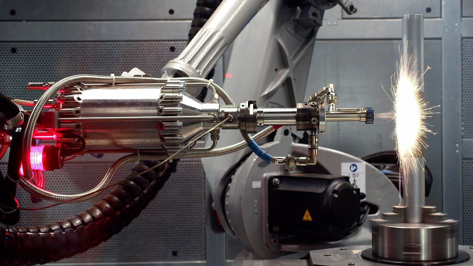 Metal robotic arm with tubes and red light spraying sparks onto a vertical metal rod in an industrial setting.