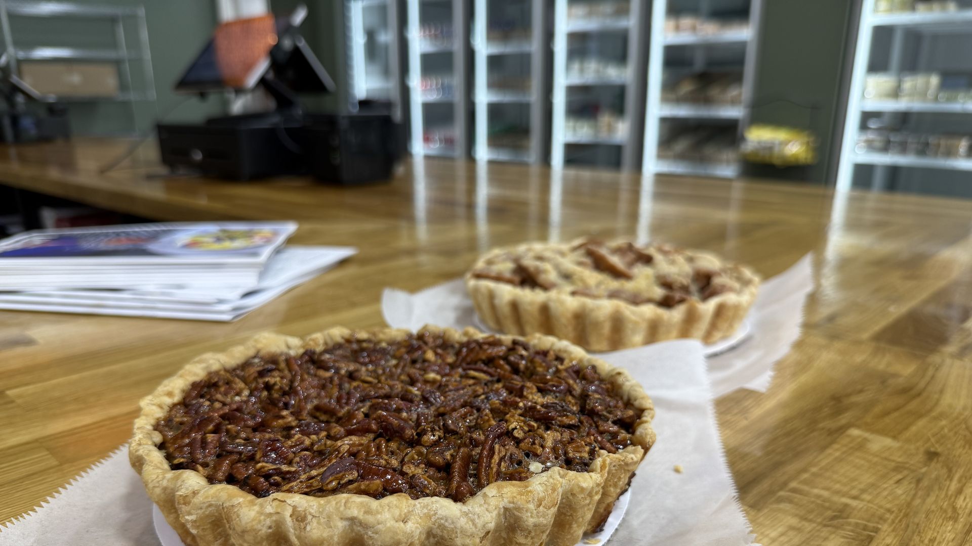 Two pecan pies on parchment paper placed on a wooden counter in a bakery setting with shelves and a cash register in the background.