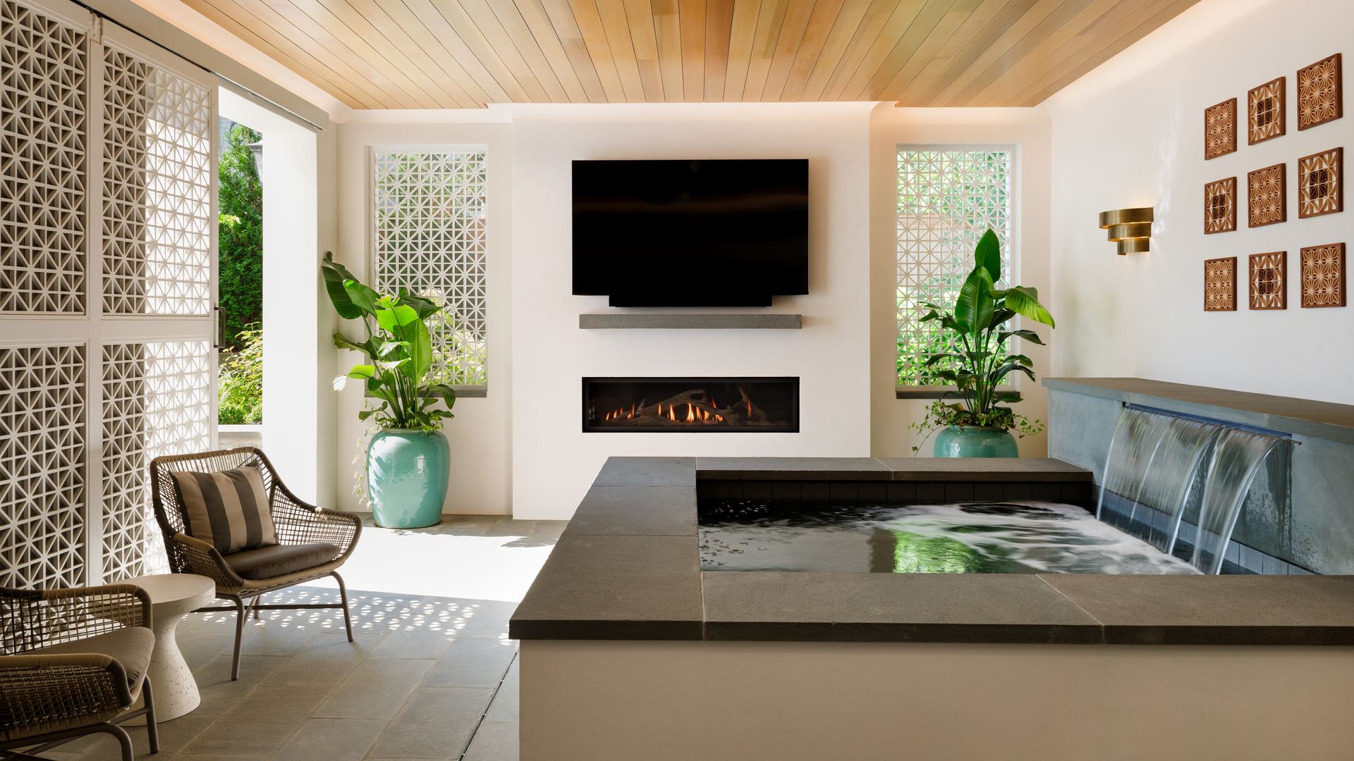 Bright indoor-outdoor lounge with a wall-mounted TV above a linear fireplace, beige ledge, and a rectangular pool to the right. There are wicker chairs, turquoise planters, lattice screens and a wood ceiling.