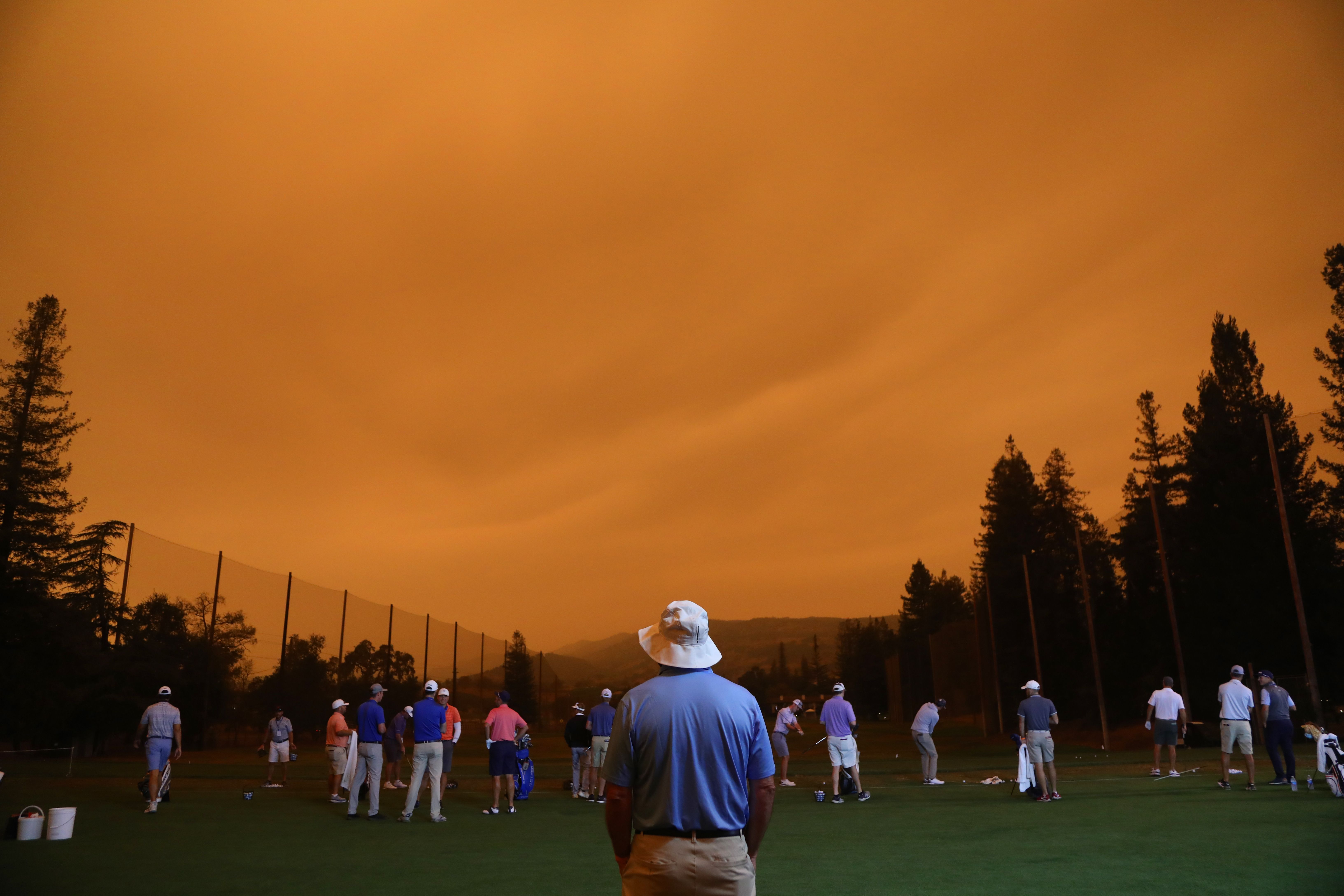 A man wearing a bucket hat stands in a field with people in the distance, under an orange sky