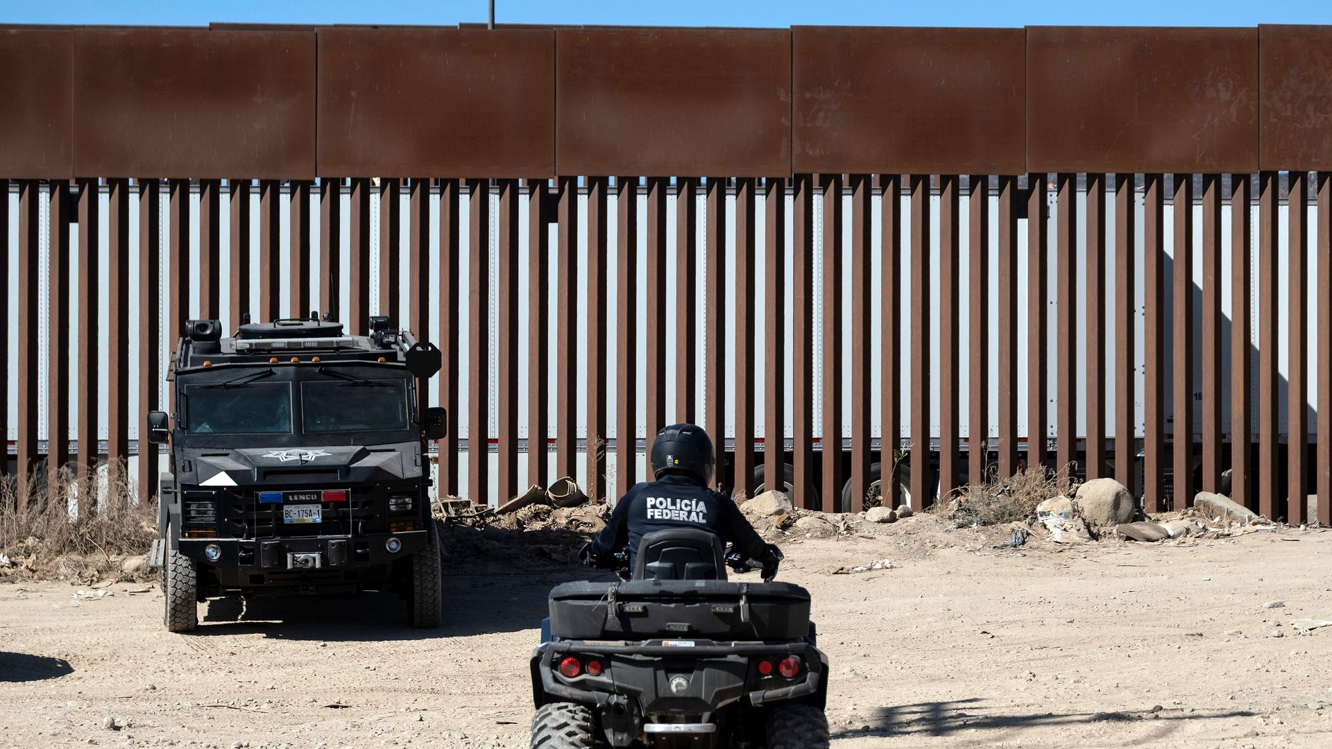 In this image, police sit in cars next to the border wall.