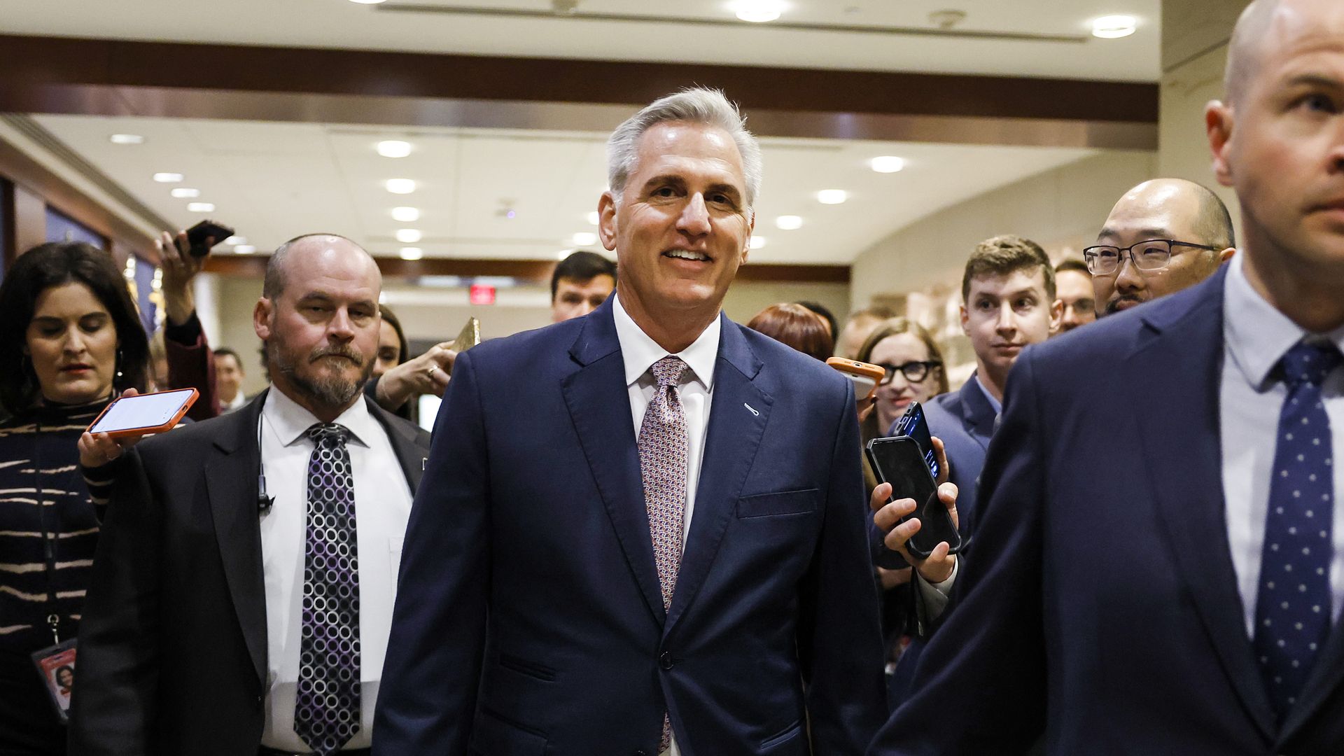 House Minority Leader Kevin McCarthy, wearing a blue suit jacket, white shirt and a red and white tie, walks in the Capitol visitors center trailed by reporters, security and staff.