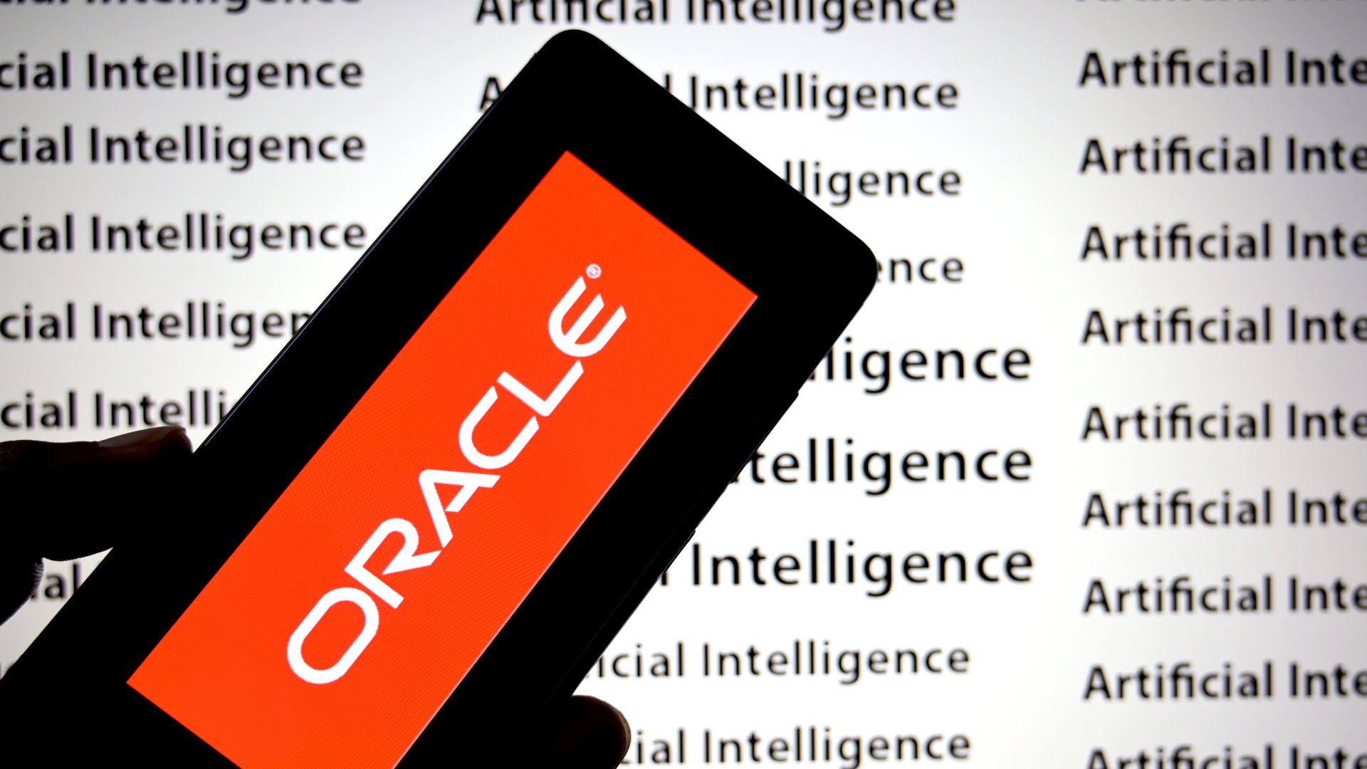 Close-up of a hand holding a black smartphone at an angle; an orange screen shows white letters that look like 'ORACLE'; the background repeats the words 'Artificial Intelligence'.