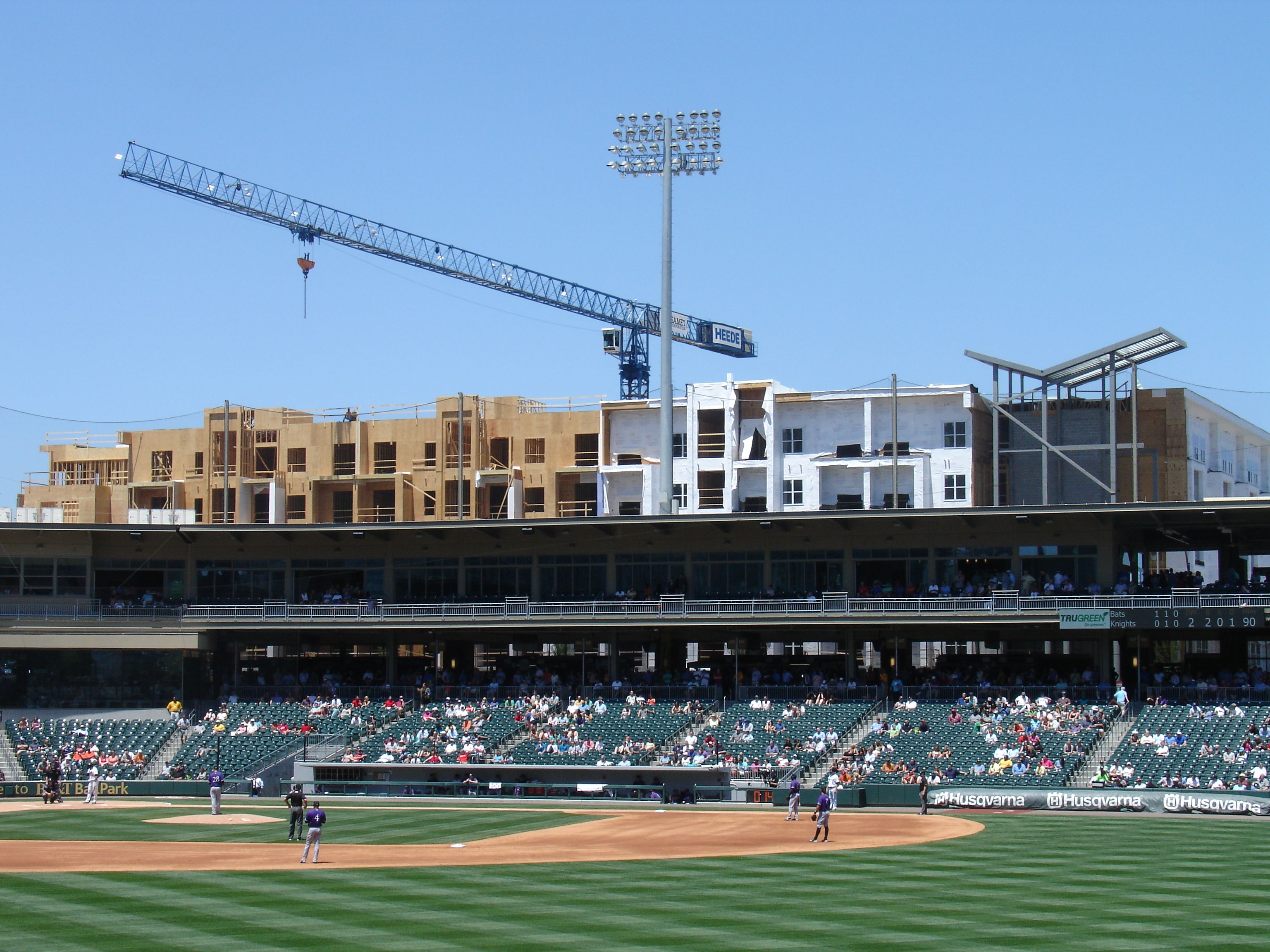 Baseball game with players in purple uniforms on a green field, spectators in stands, and an under-construction beige and white building with a blue crane behind the stadium under a clear blue sky.