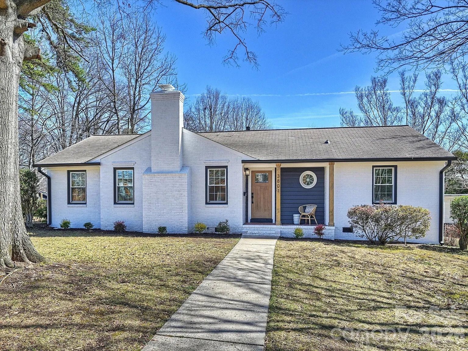 Single-story white brick house with a central chimney, wooden front door, navy blue siding on porch, round window, and a chair, under a clear blue sky with leafless trees.