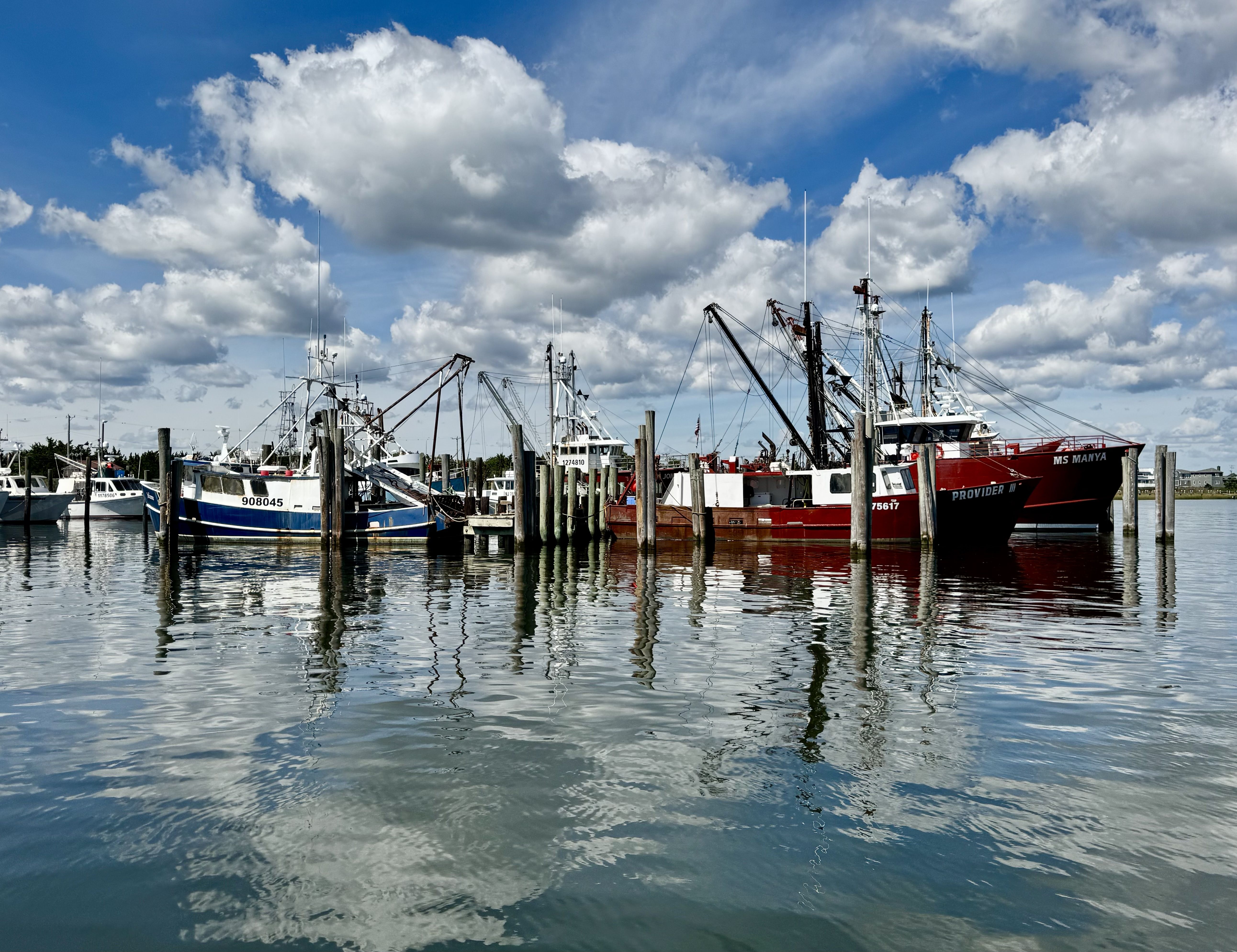 A marina scene with several fishing boats docked at wooden piers, tall masts and rigging against a bright blue sky filled with fluffy white clouds; calm water reflects the vessels.