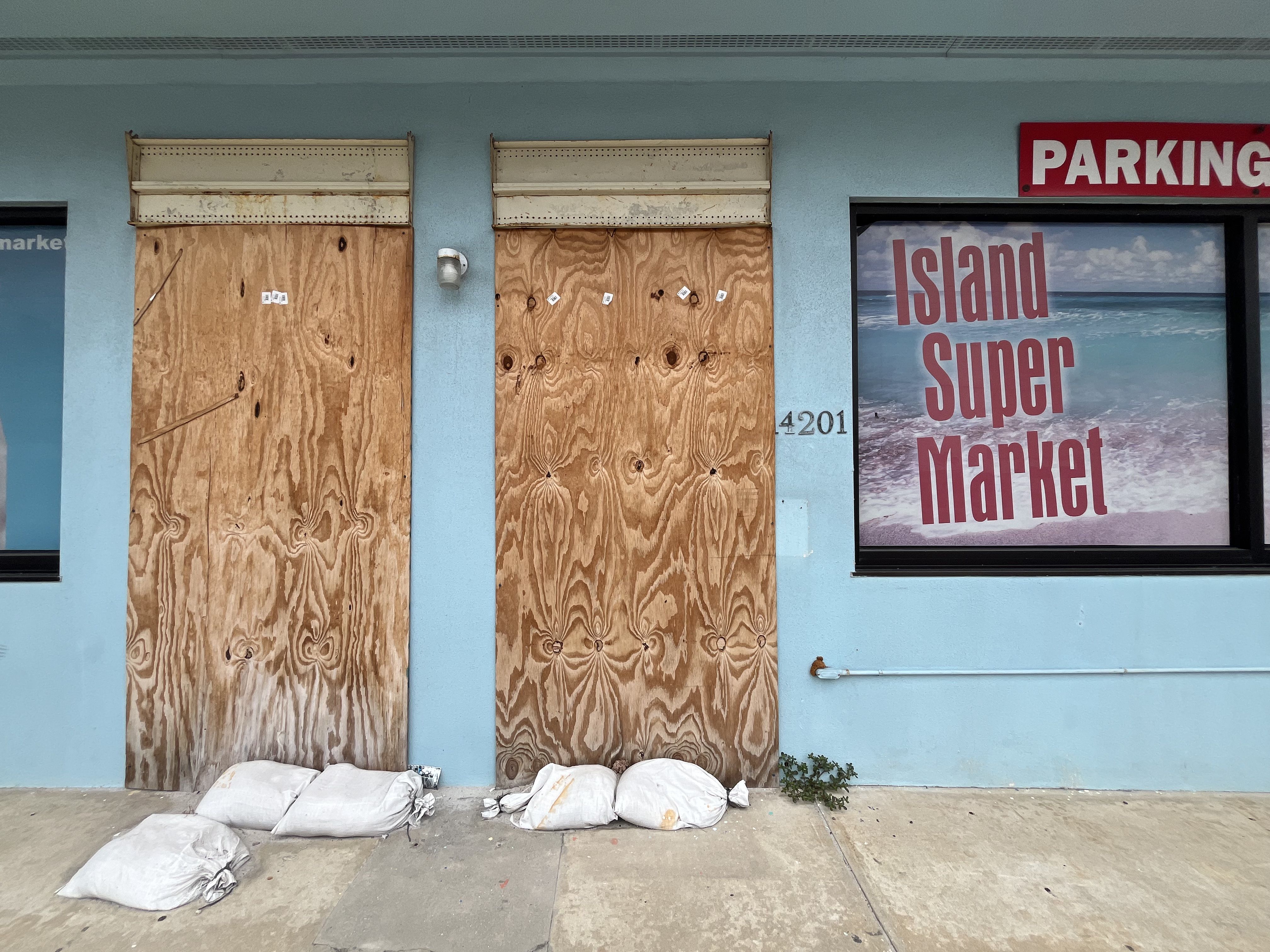 Light blue storefront with two plywood-covered doors and sandbags at the base, next to a window sign reading "Island Super Market" with an ocean beach background.
