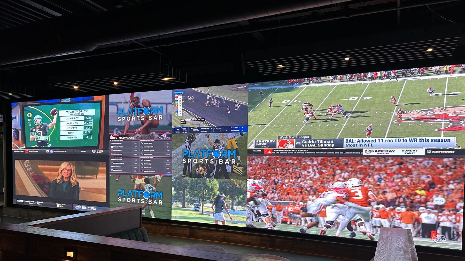 Wall of several large screens in a sports bar, showing mixed sports content: a football game on a green gridiron, branding that reads "Platform Sports Bar", and other channel graphics; dark ceiling and a wooden counter in the foreground.