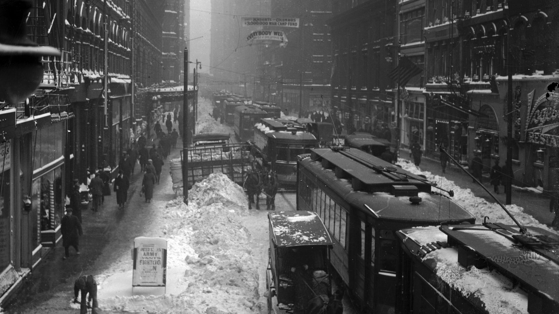 Black-and-white photo of a snow-covered city street with horse-drawn carriages, streetcars, and pedestrians amid tall buildings and snow piled along the road.