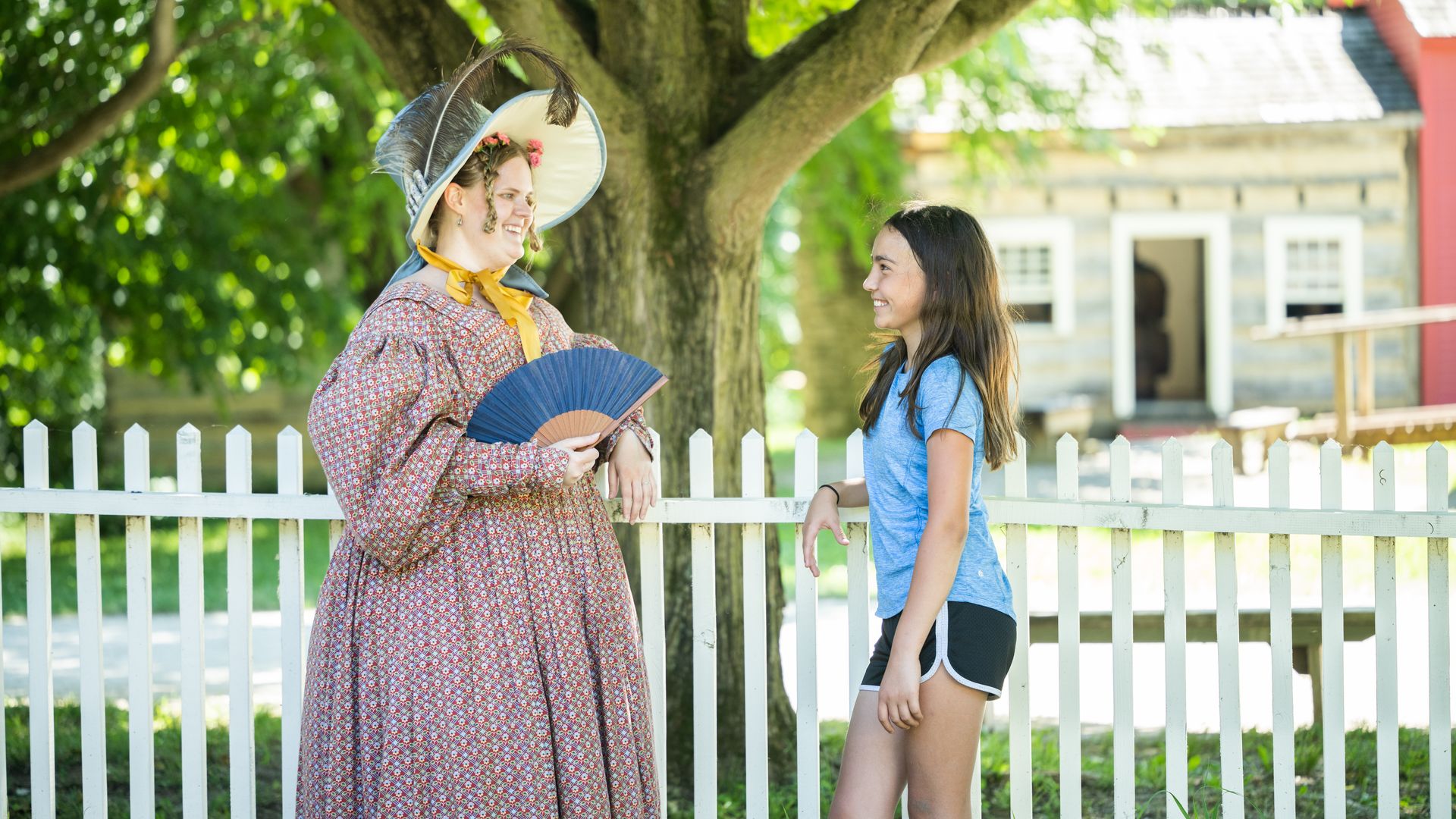 A Conner Prairie interpreter in period clothing chats with a visitor in front of a white fence.