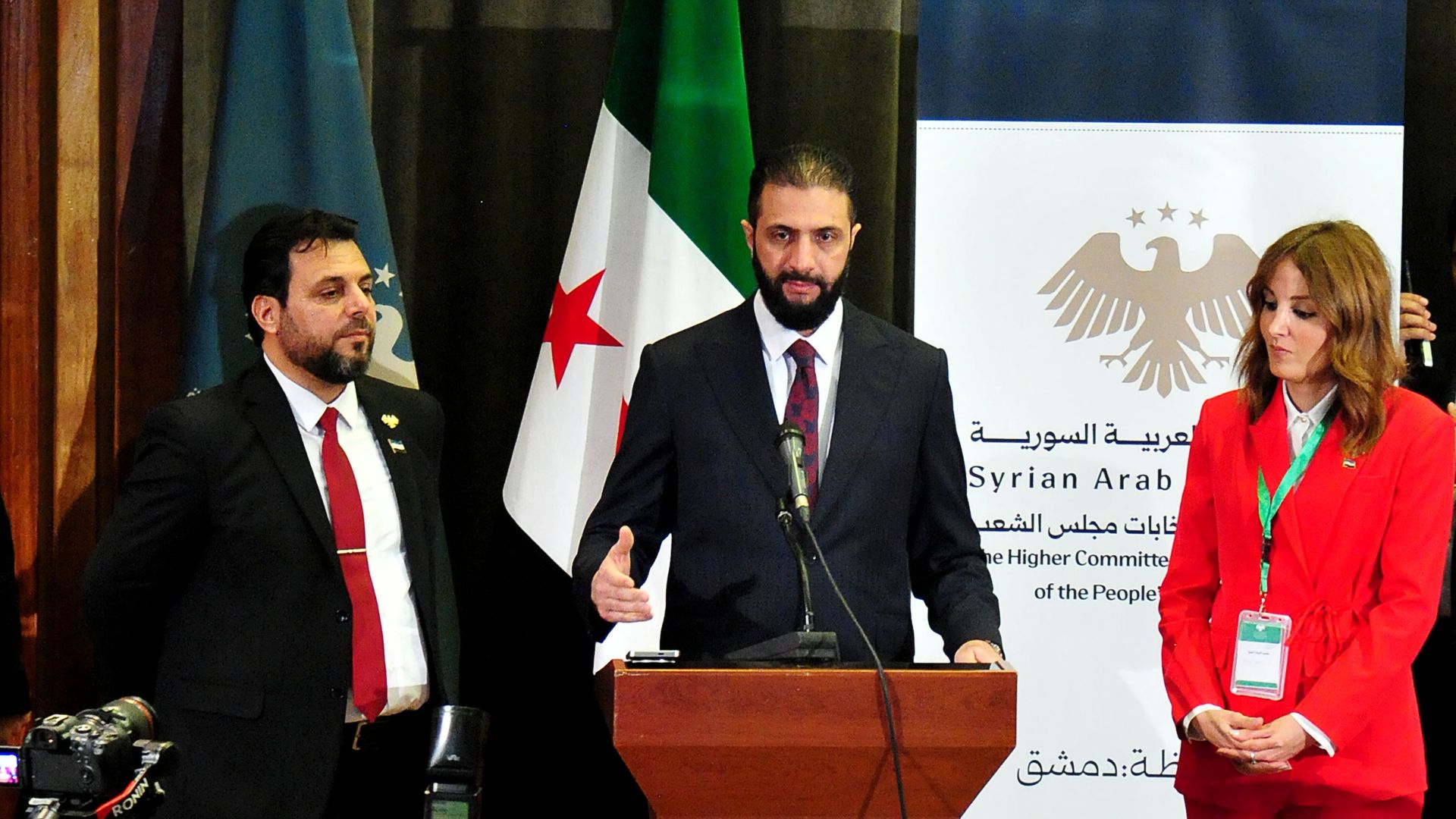 Man at podium with woman in red suit at his side and man in suit at his other side
