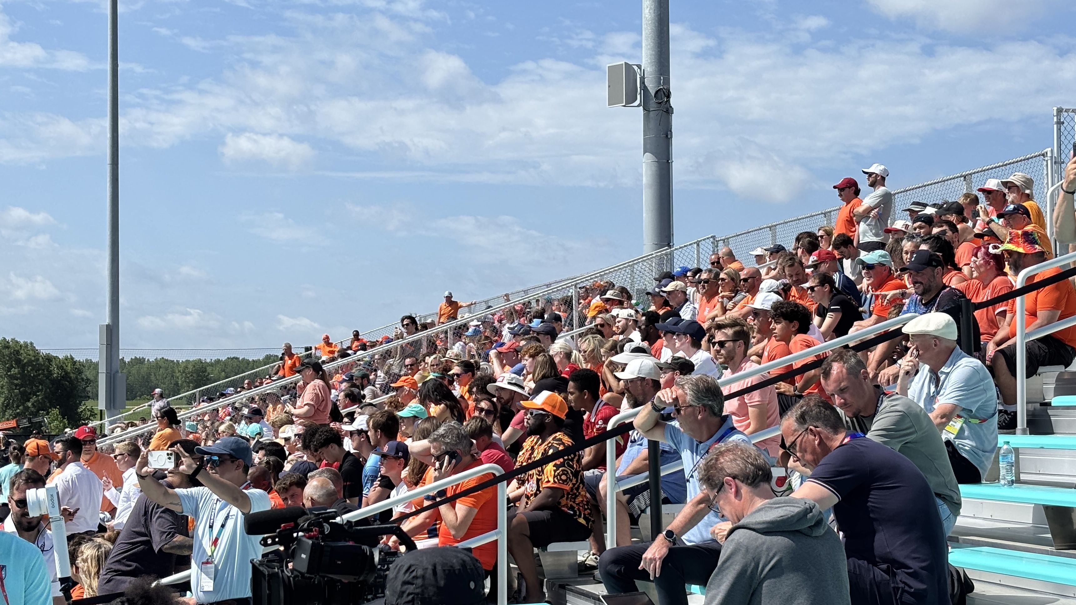 Crowd of spectators in stadium stands under a blue sky, many wearing orange shirts. A chain-link fence runs along the seating; a camera crew with a large camera sits at the front.