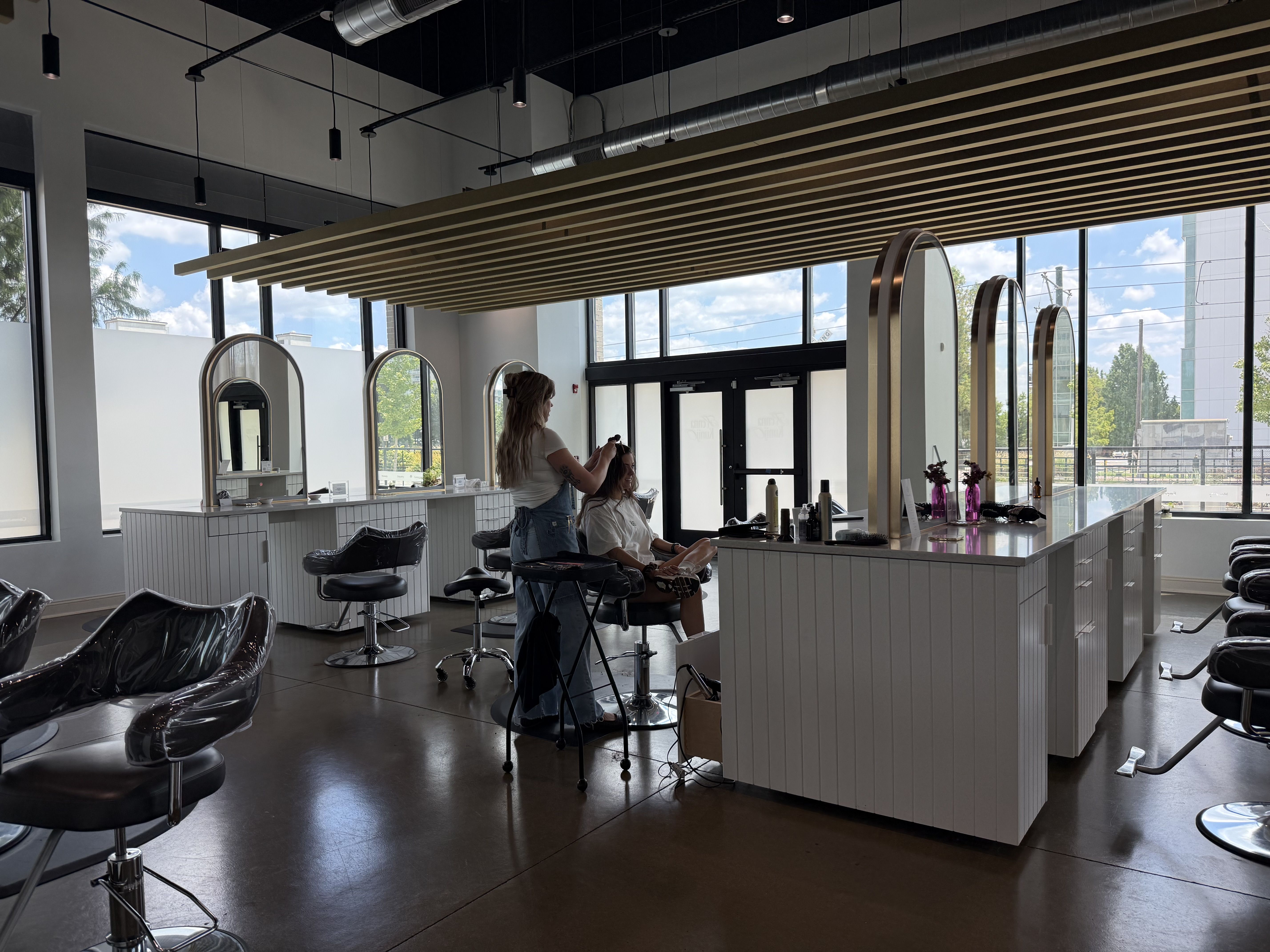 Sunlit modern hair salon with large windows, copper-framed arched mirrors, and white tiled counters. A stylist works on a client's hair at a chair beneath a slatted wood ceiling with exposed ducts.