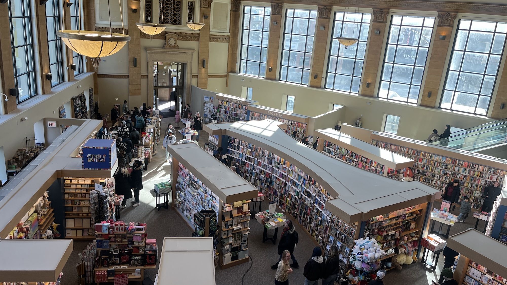 Interior of a large, sunlit bookstore with tall windows, wooden accents, shelves filled with books and merchandise. People browse and chat beside labeled sections like Favorites and Fiction.