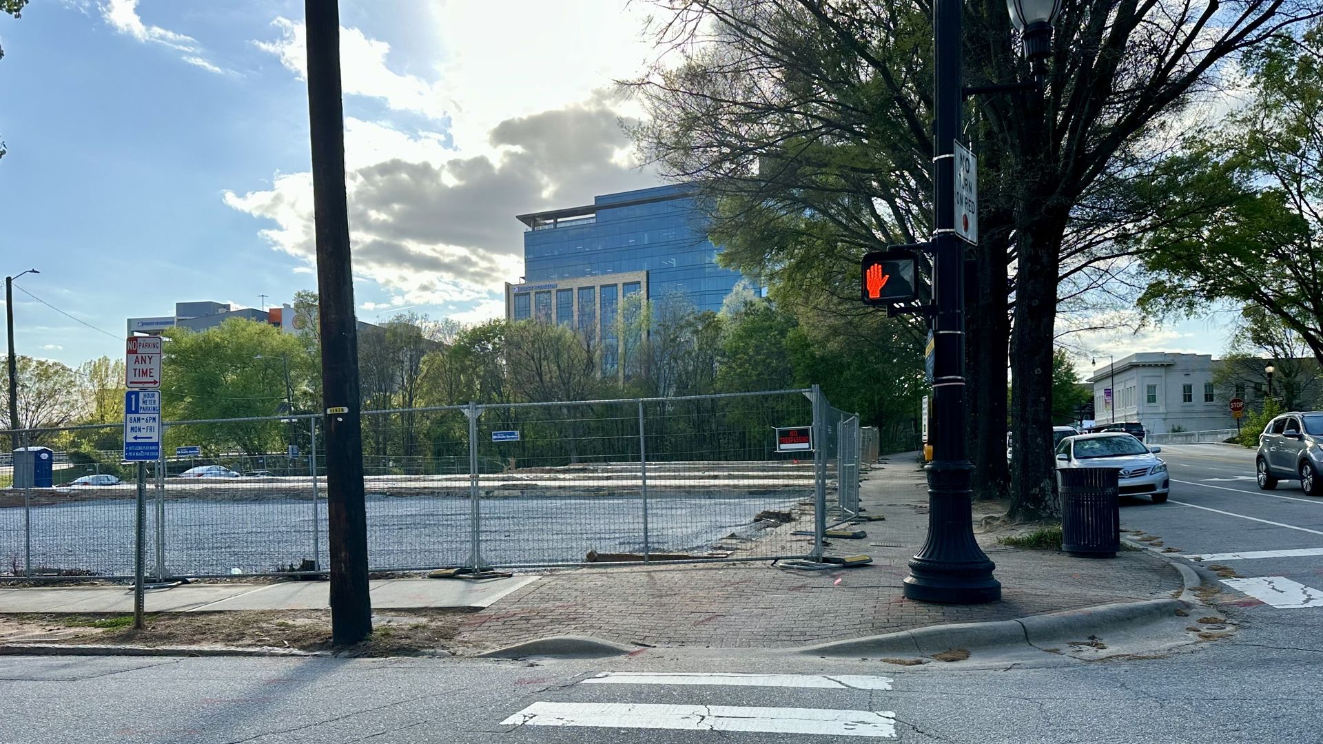 Crosswalk on a city corner with cracked pavement, a tall black lamppost showing a red don't-walk signal, chain-link fencing along a waterfront, and a blue glass office building in the background with trees and cars.