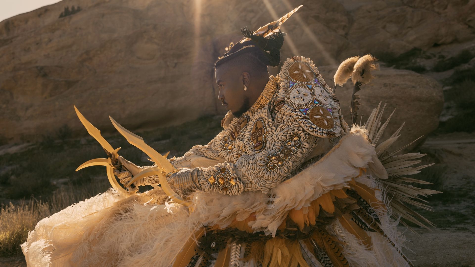 Chief Adjuah, wearing an ivory and gold beated suit with feathers, brandishes two weapons as the sun flares down.